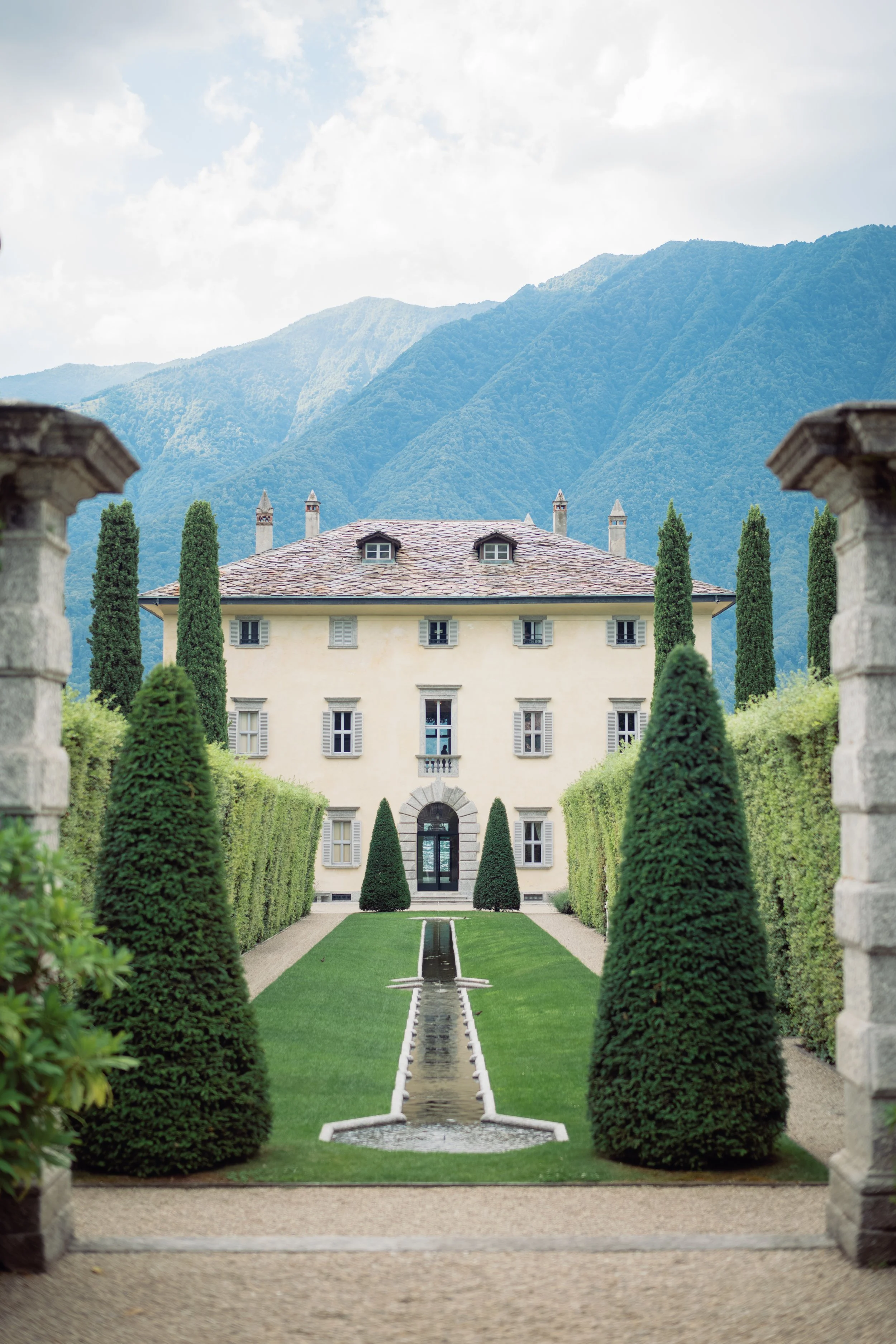 A symmetrical garden with a fountain running along a path, flanked by tall, manicured cypress trees leading to a historic villa with a tiled roof, set against green mountains under a cloudy sky.