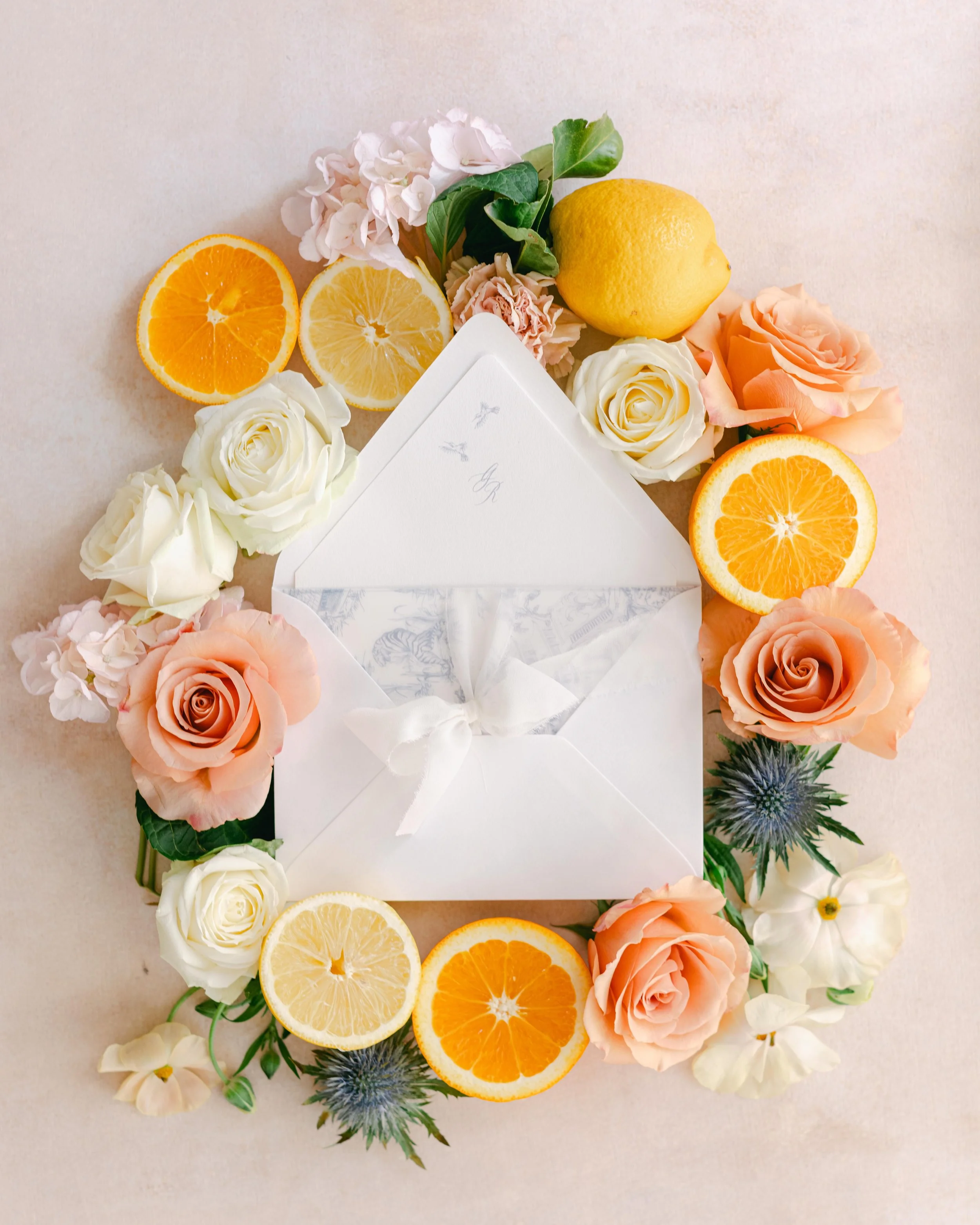 A floral arrangement with white, cream, and peach roses, pink hydrangeas, a lemon, and orange slices surrounding an open white card with cursive writing.