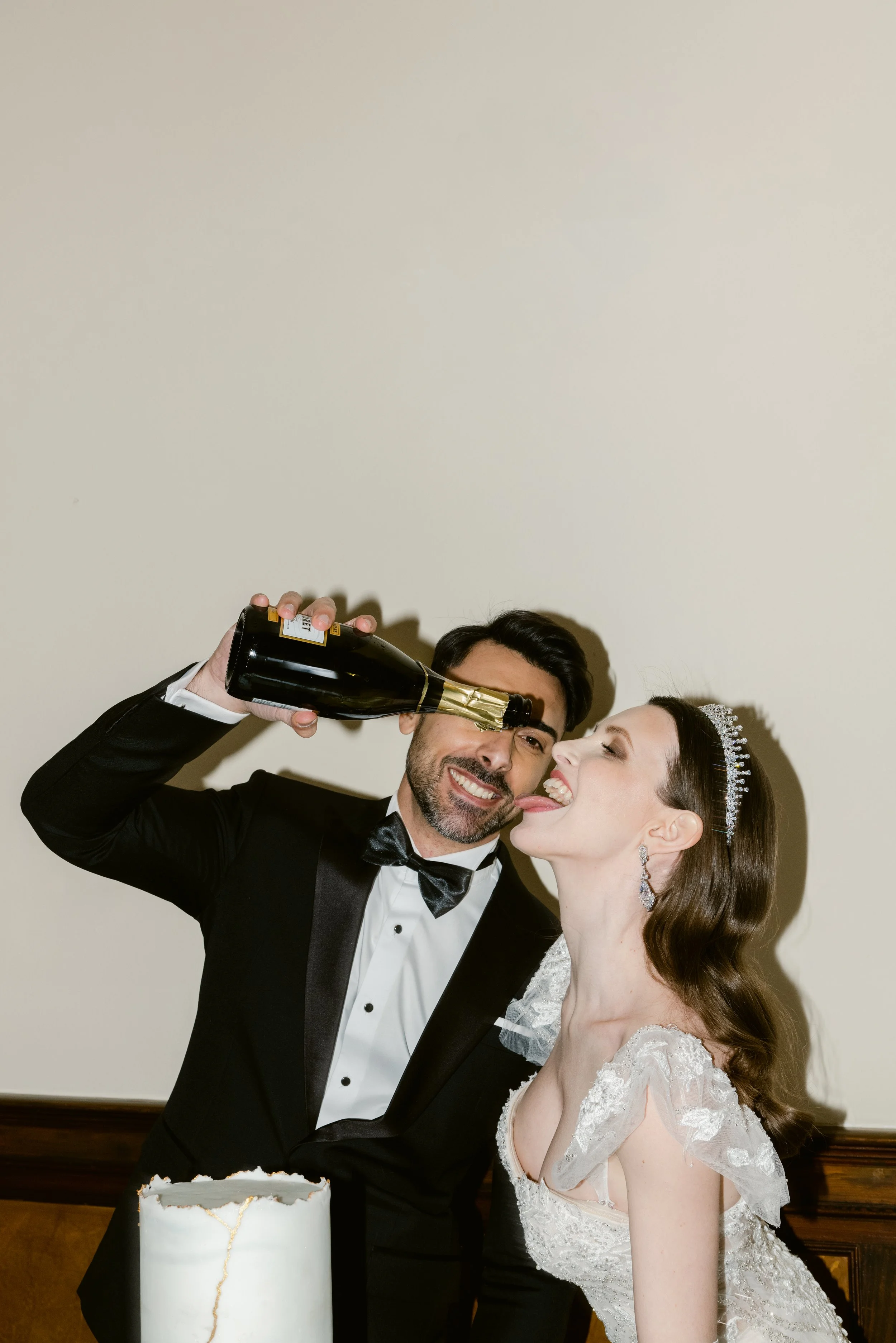 A man in a tuxedo pouring champagne into the mouth of a woman in a wedding dress, who is smiling with her tongue out. They are celebrating with a white wedding cake in front of them.