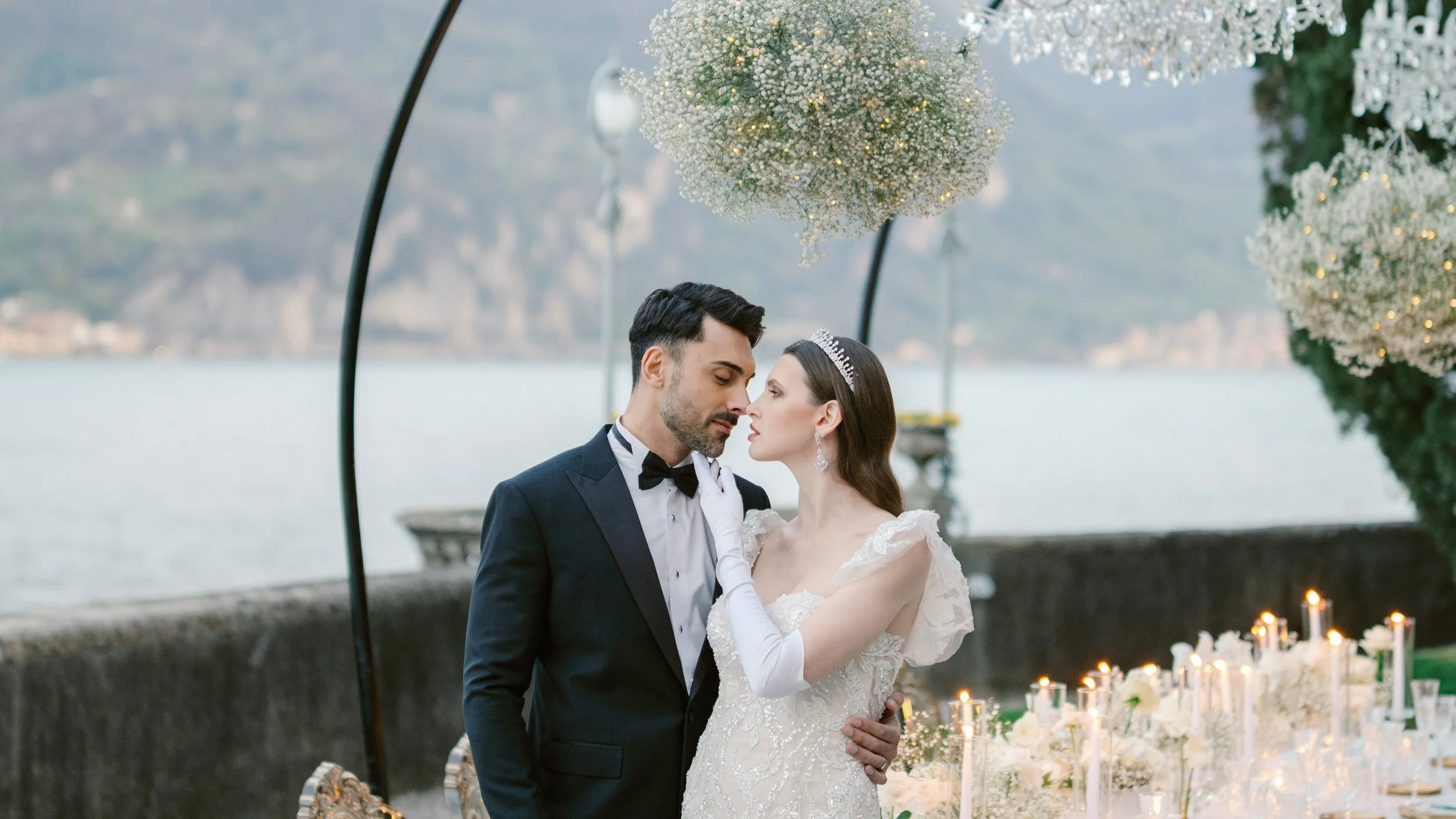 A bride and groom stand close together under an umbrella decorated with white flowers and lights, near a body of water with mountains in the background, during a wedding ceremony.