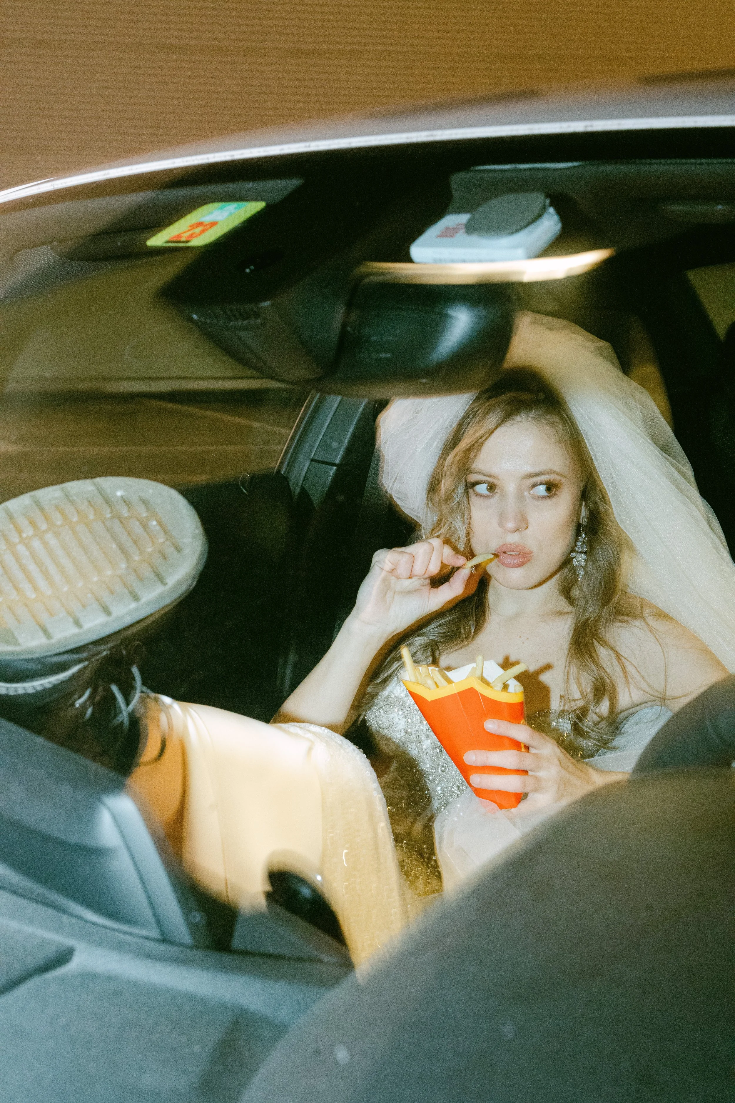 A young woman in a wedding dress and veil, sitting in a car, eating fries from a red container.