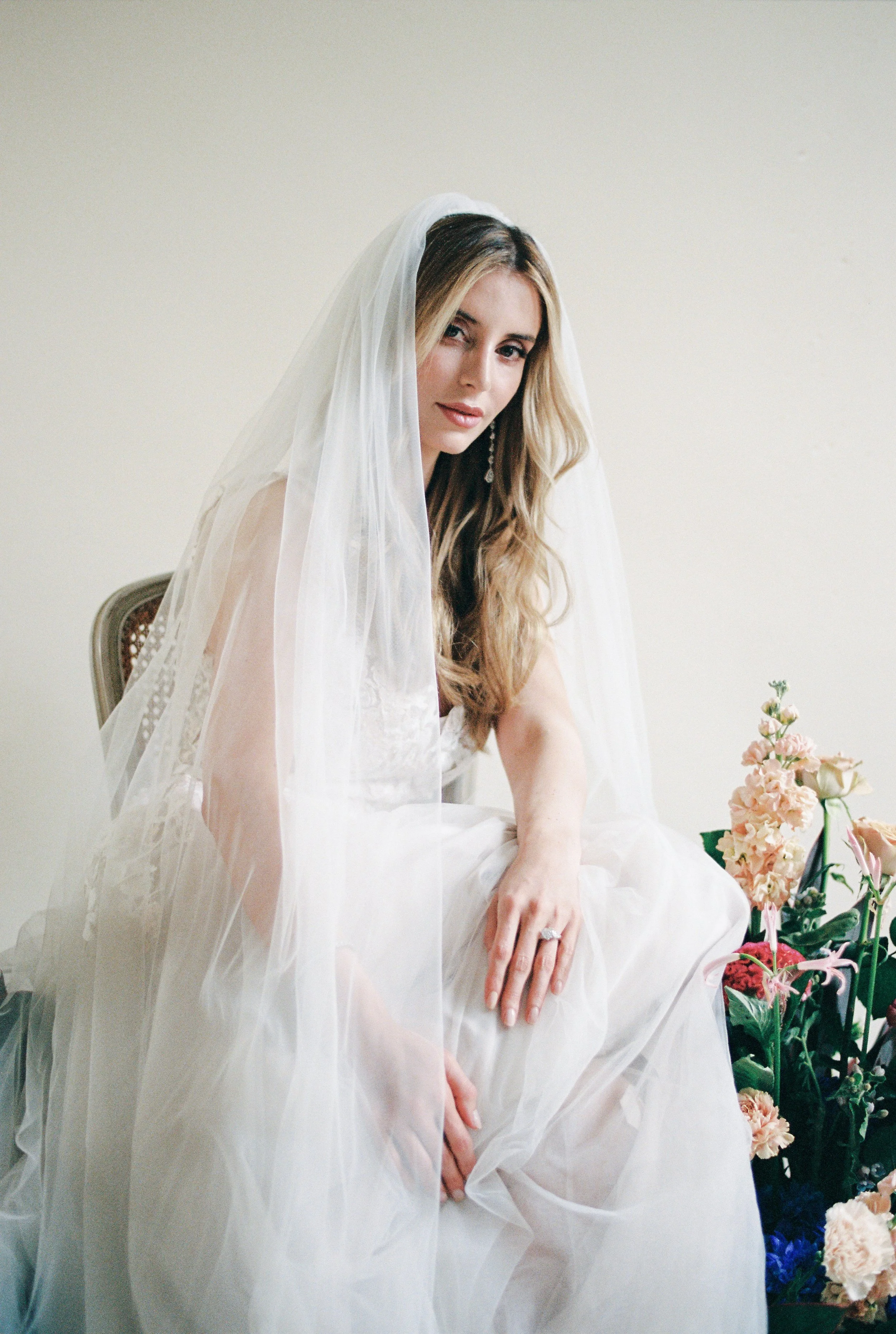 A woman in a wedding dress with a white veil, sitting next to a bouquet of pink and white flowers against a plain background.