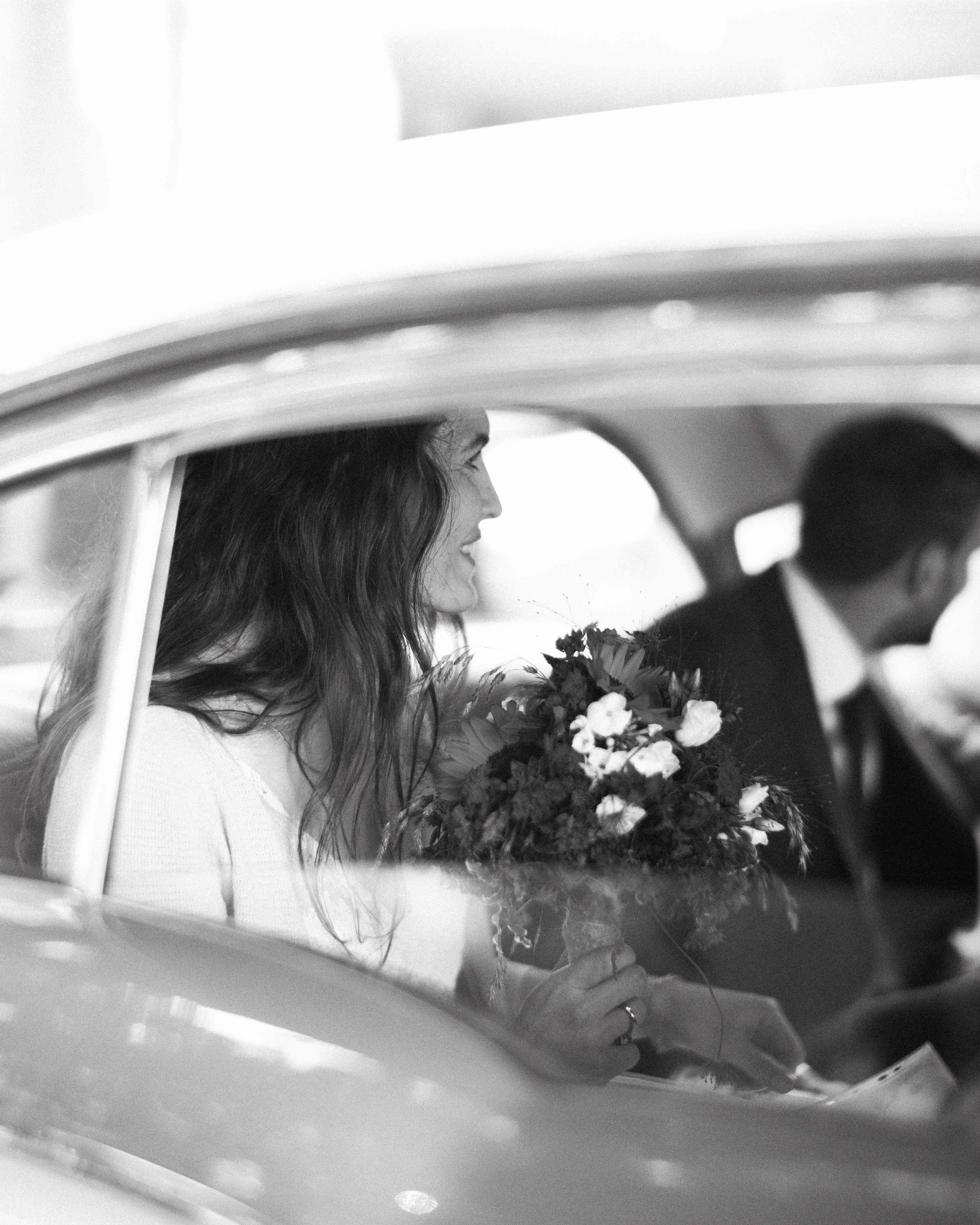 A woman with long hair sitting inside a vintage car holding a bouquet of flowers. The photo is in black and white.