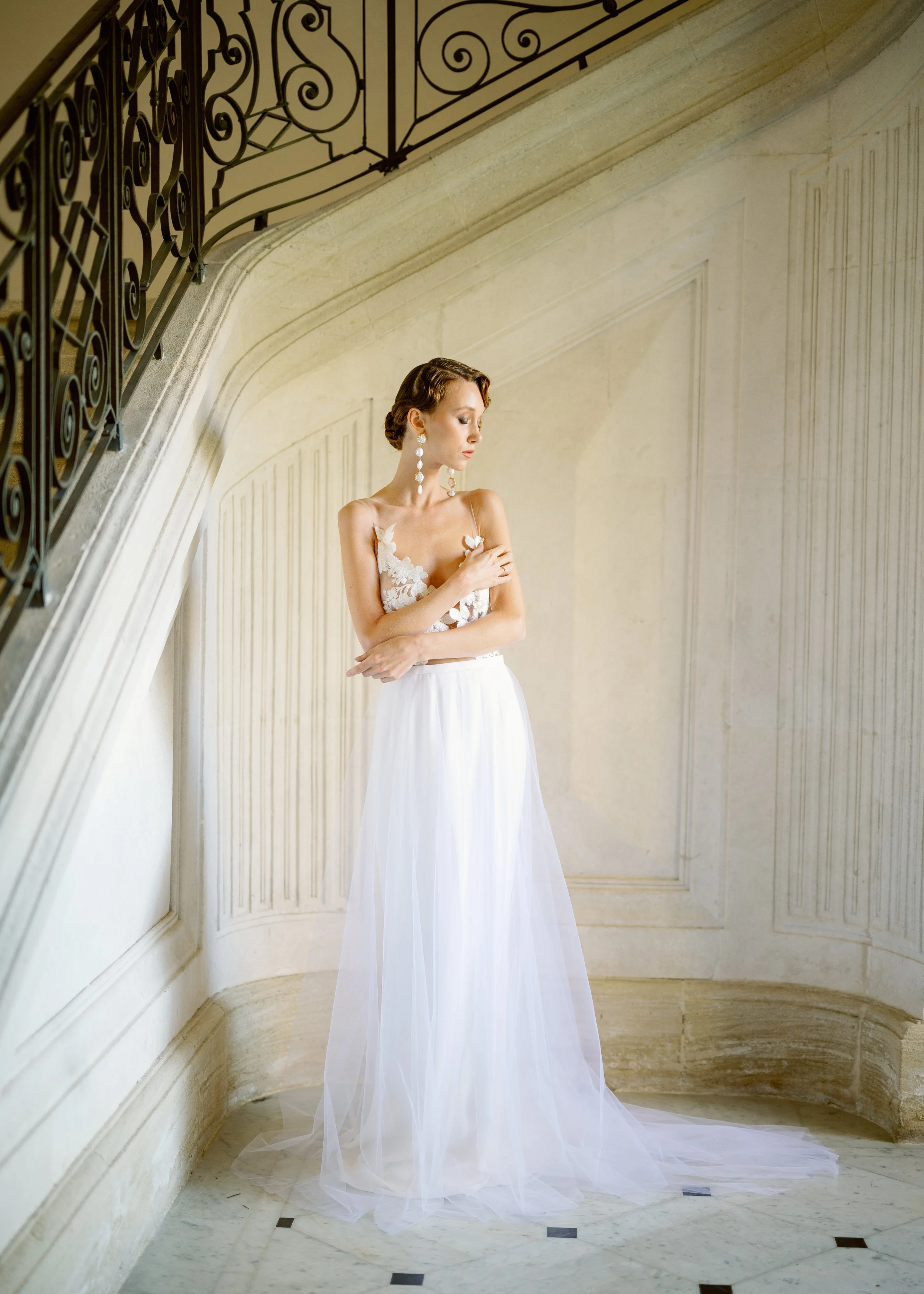 A woman in a white wedding dress with floral lace details on the bodice, standing under a curved staircase with a decorative iron railing, in a beige-toned interior with marble flooring. She has short, styled hair and wears long, elegant earrings, wi