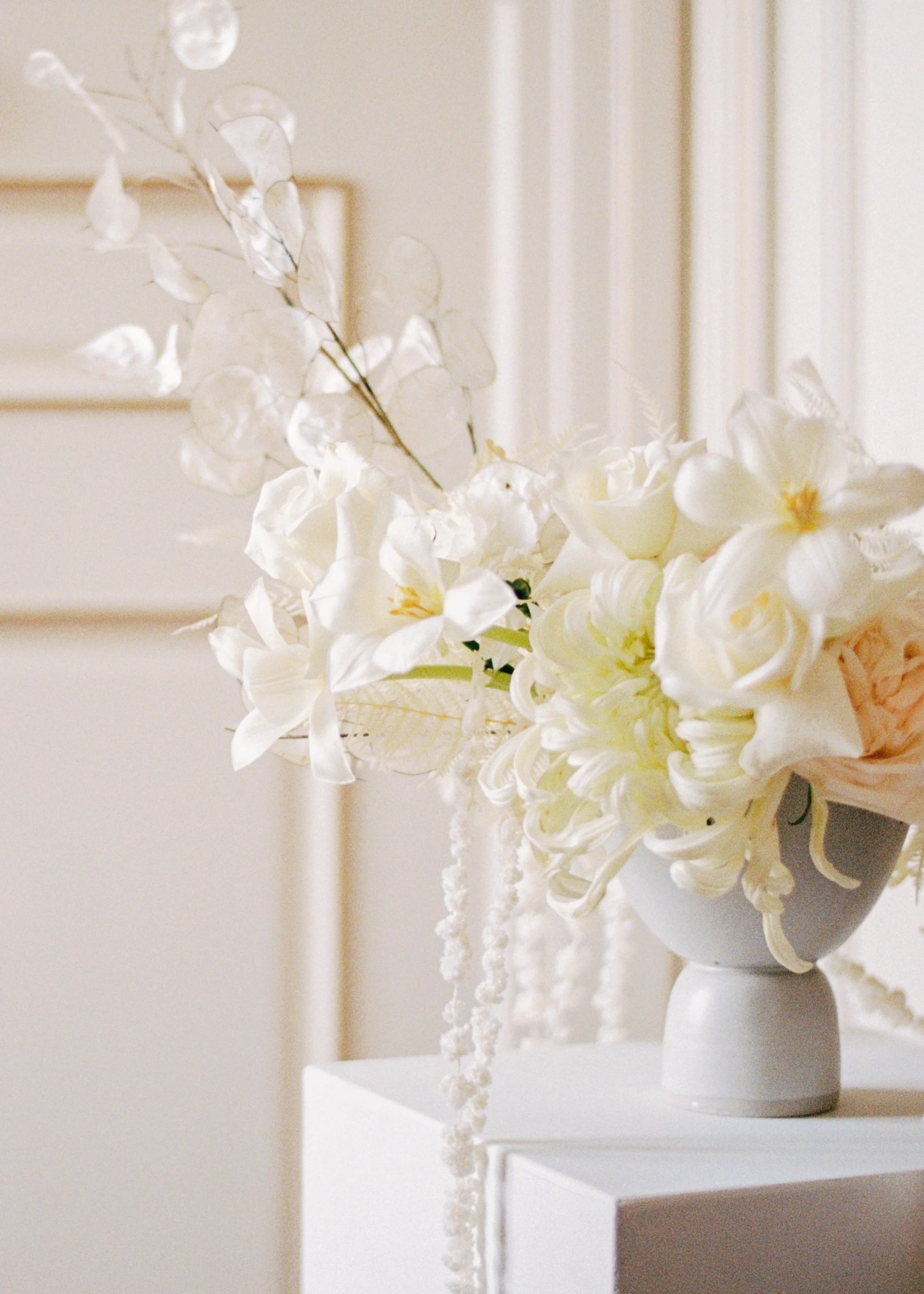 A white vase on a white pedestal holding a bouquet of white flowers and white dried botanical elements, with a neutral beige wall and framed art in the background.