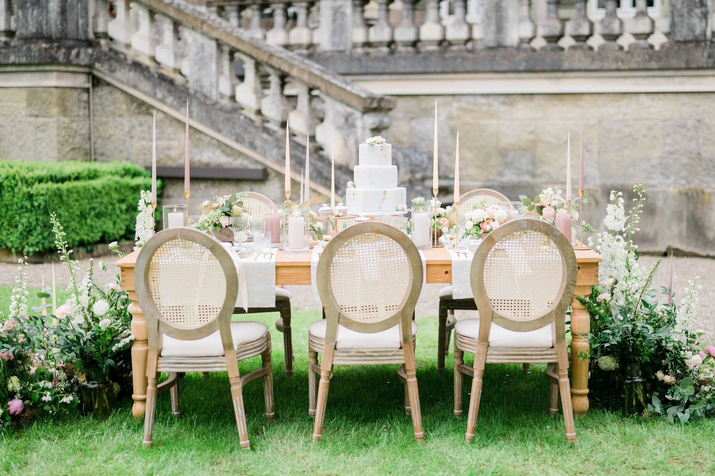 An outdoor table set for a celebration with a white tiered cake and surrounded by chairs and floral arrangements, with stone stairs and a stone wall in the background.