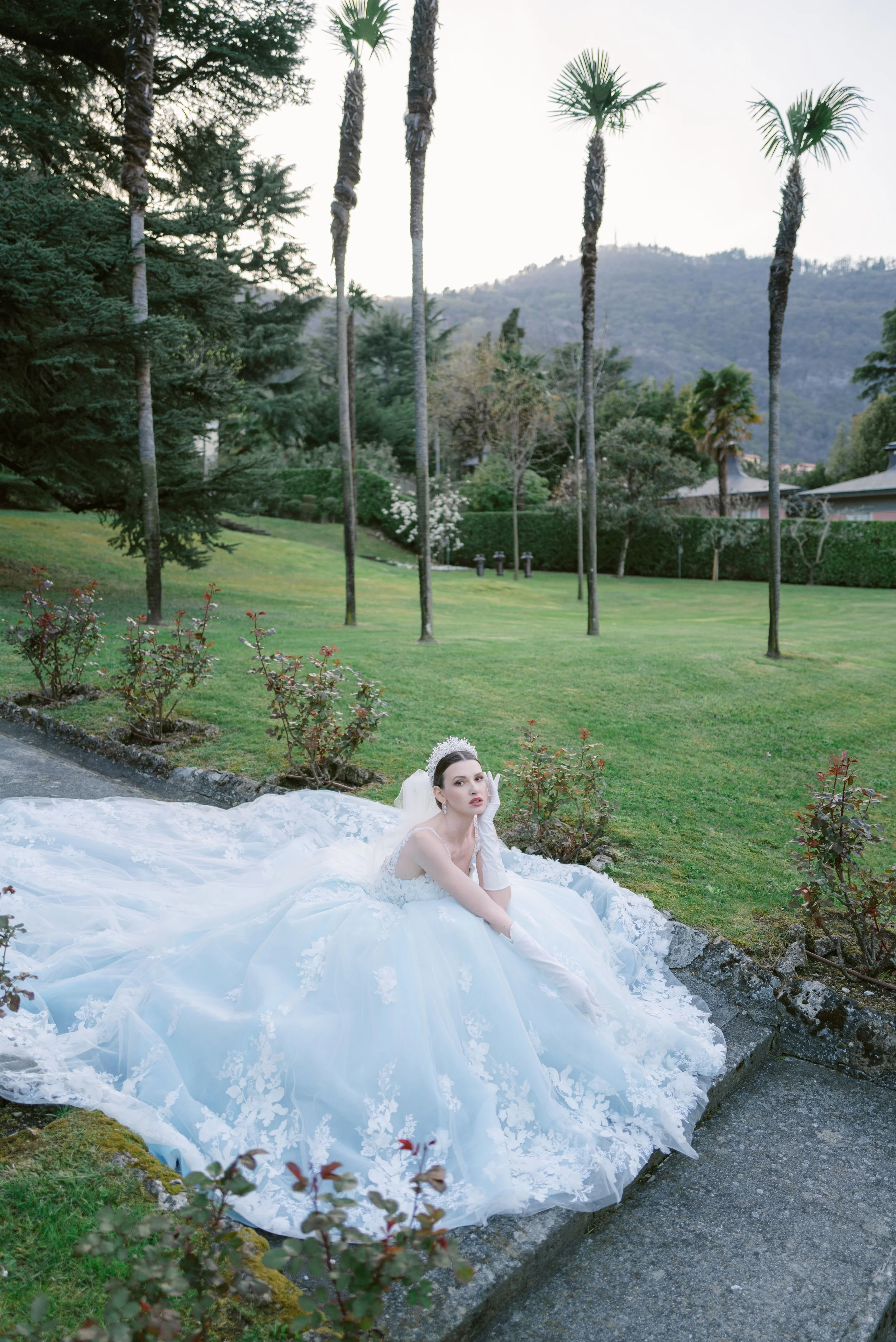 A woman in a white wedding gown sitting on the ground outdoors in a garden with green grass, trees, and palm trees. She is wearing white gloves and has her hair styled in an updo, with a serene expression on her face.