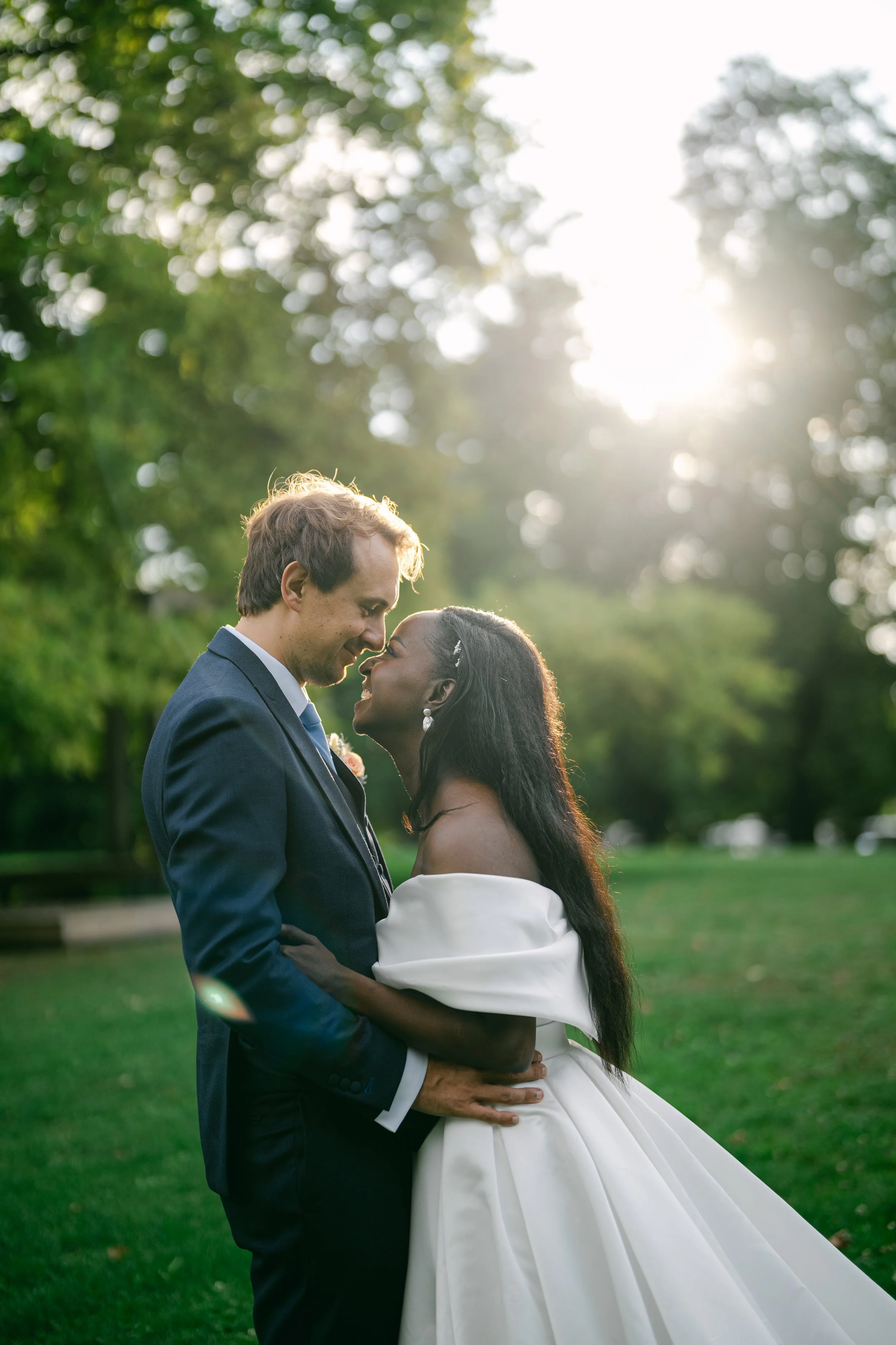 A newlywed couple, a man in a dark suit and a woman in a white wedding dress, standing close together outdoors in a park with trees and green grass, facing each other with foreheads touching, during sunset with sunlight filtering through the trees.