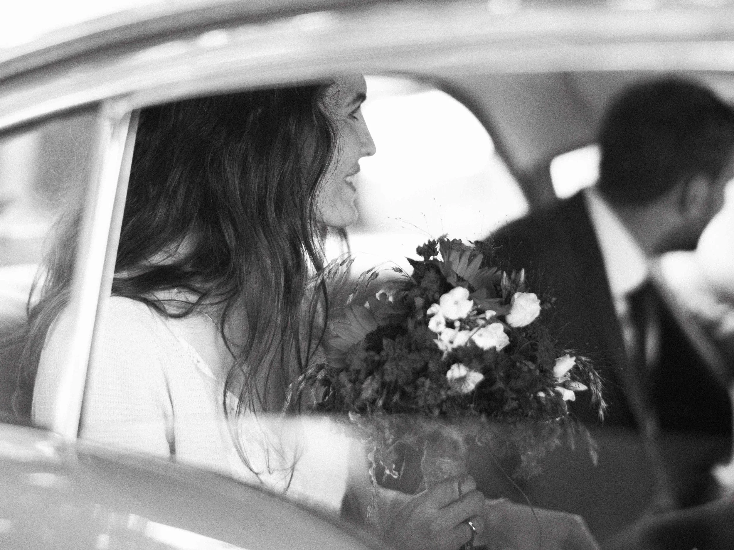 Black and white photo of a woman with long hair holding a bouquet of flowers inside a car, with a man in the driver's seat.