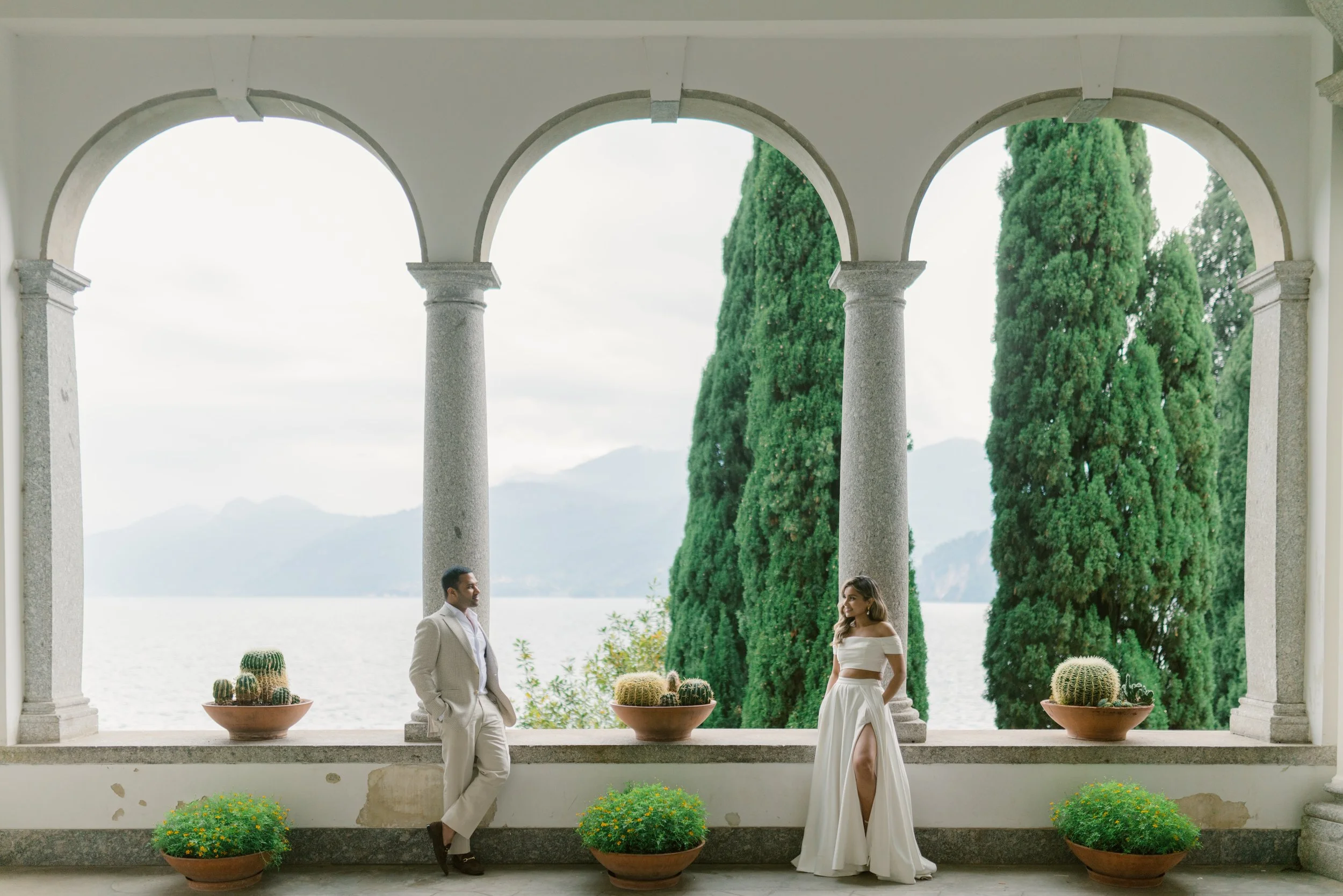 A couple dressed in white standing and talking on a balcony with potted cacti and marble columns, overlooking a landscape with tall trees and mountains in the distance.