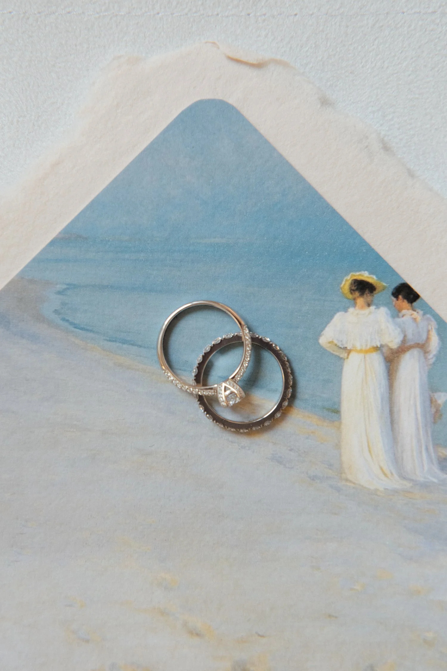 Close-up of two rings, one with small diamonds and one with black stones, resting on a vintage postcard featuring an illustration of two women in white dresses with wide yellow hats, standing on a boat with a large mountain in the background.