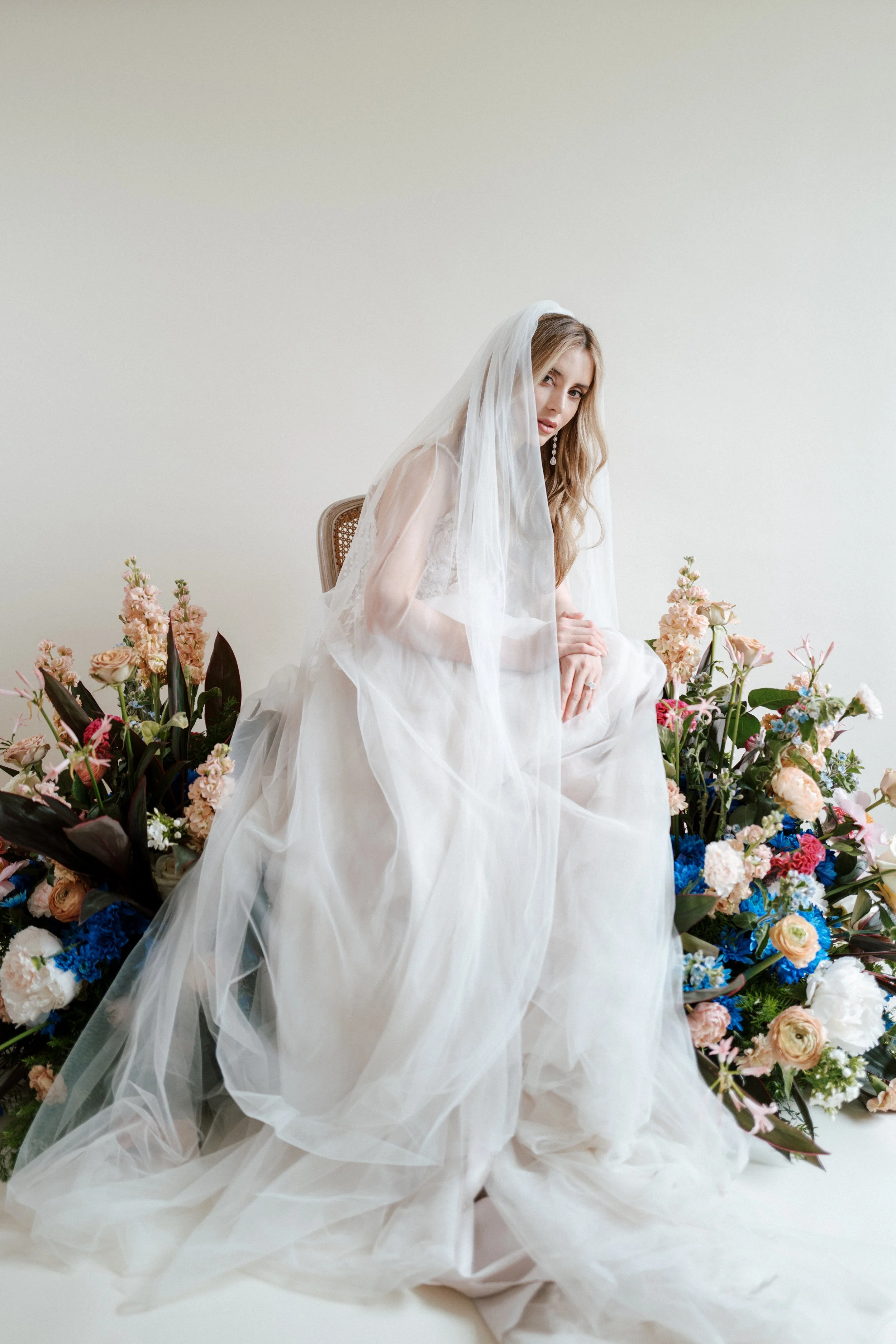 A woman wearing a white wedding dress and veil seated amidst a large colorful flower arrangement against a plain light-colored background.