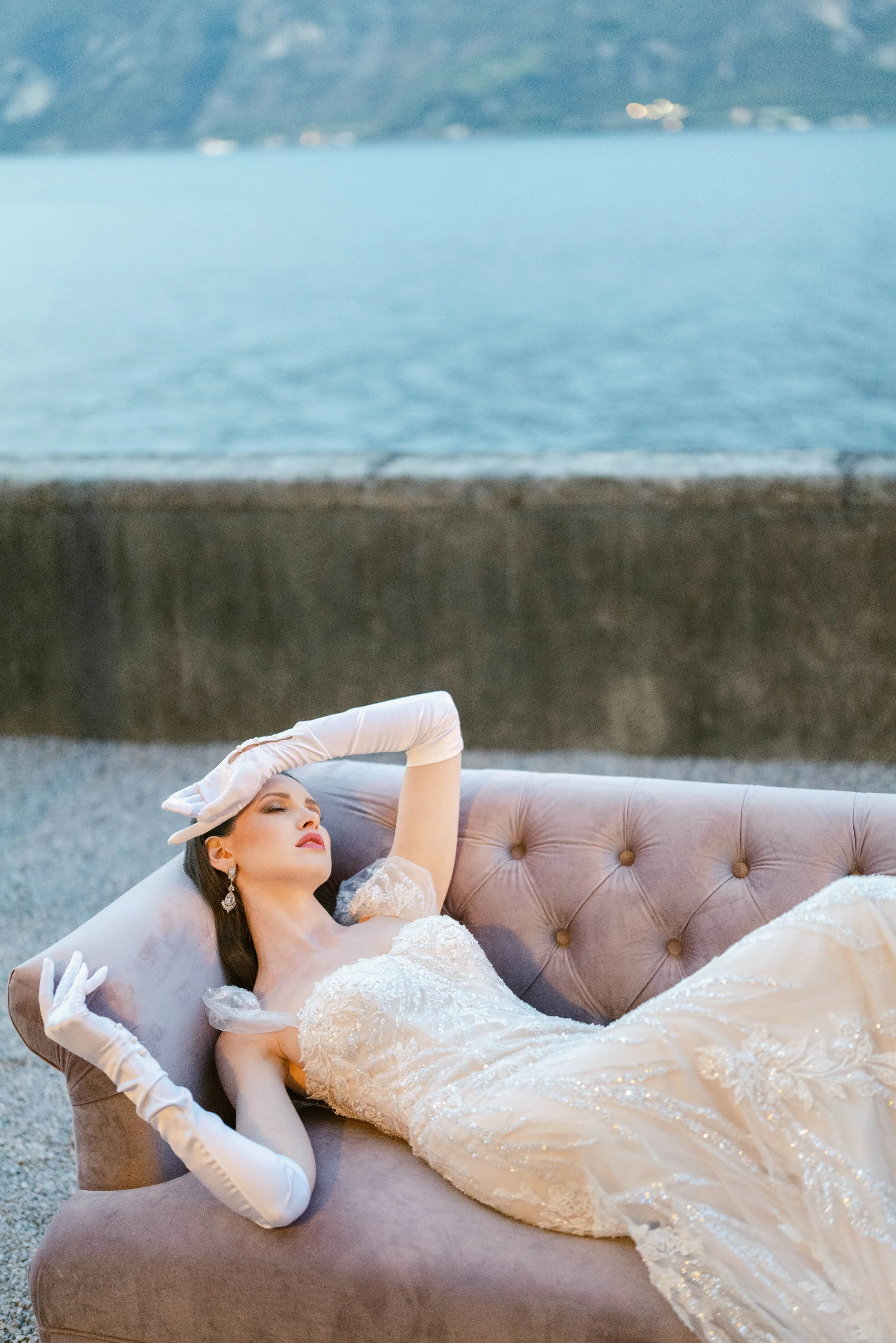 A woman in an elegant white wedding dress is lying on a plush beige sofa outdoors, eyes closed, with her left arm resting on her forehead, wearing white gloves and earrings. The background includes a body of water and distant mountains.