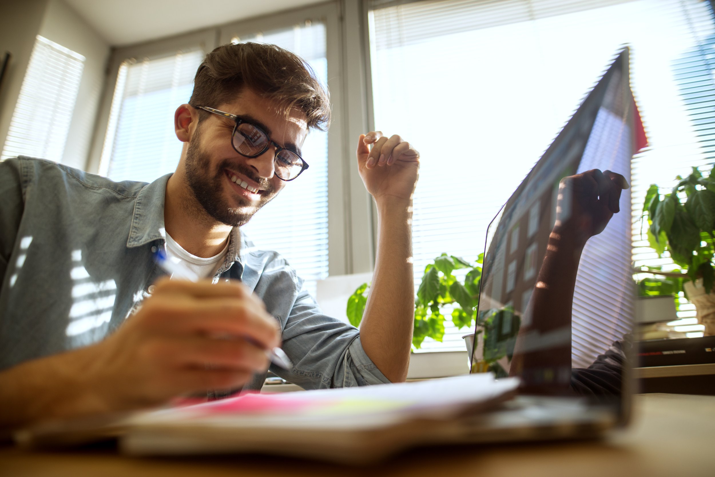 A smiling man wearing glasses works at a desk with a laptop and notebooks, sitting in a bright office with large windows and plants.