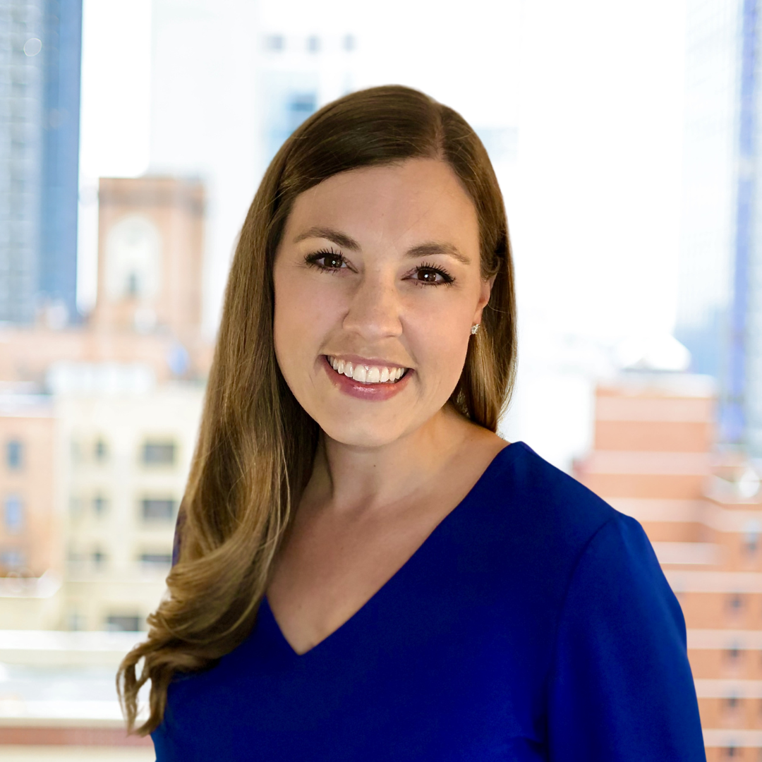 A woman with long brown hair wearing a blue top, smiling in front of a blurred cityscape background.