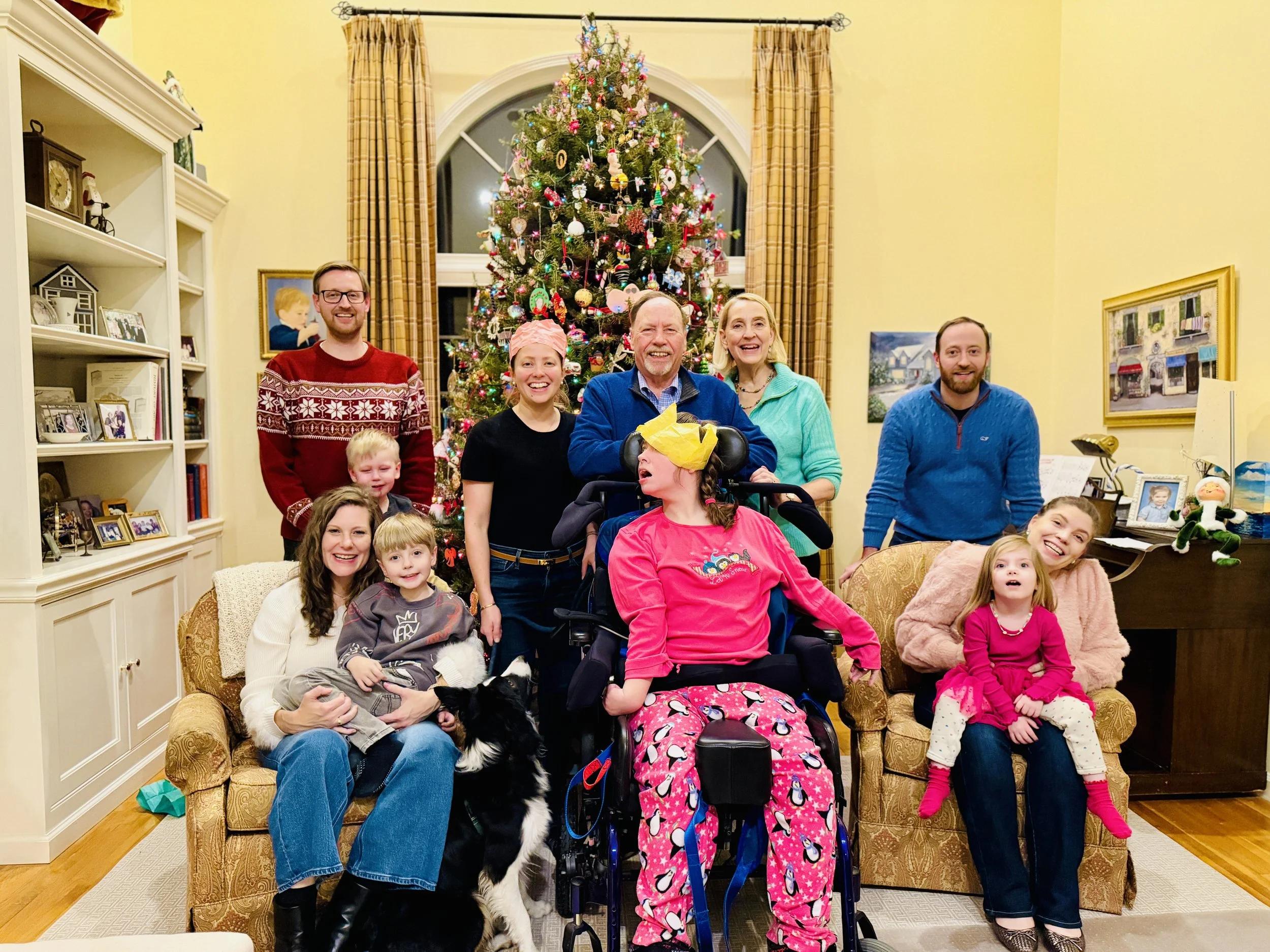 Family gathering in living room with decorated Christmas tree, smiling group of adults and children, some sitting on a couch and others standing, in festive sweaters and pajamas.