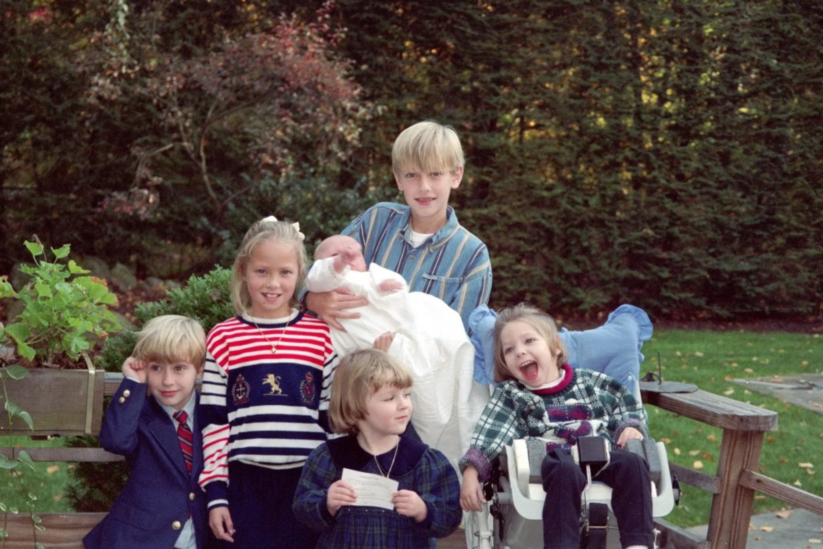 A group of six children and a baby posing outdoors in front of a backdrop of trees and bushes. The children are smiling and appear to be enjoying a family gathering.