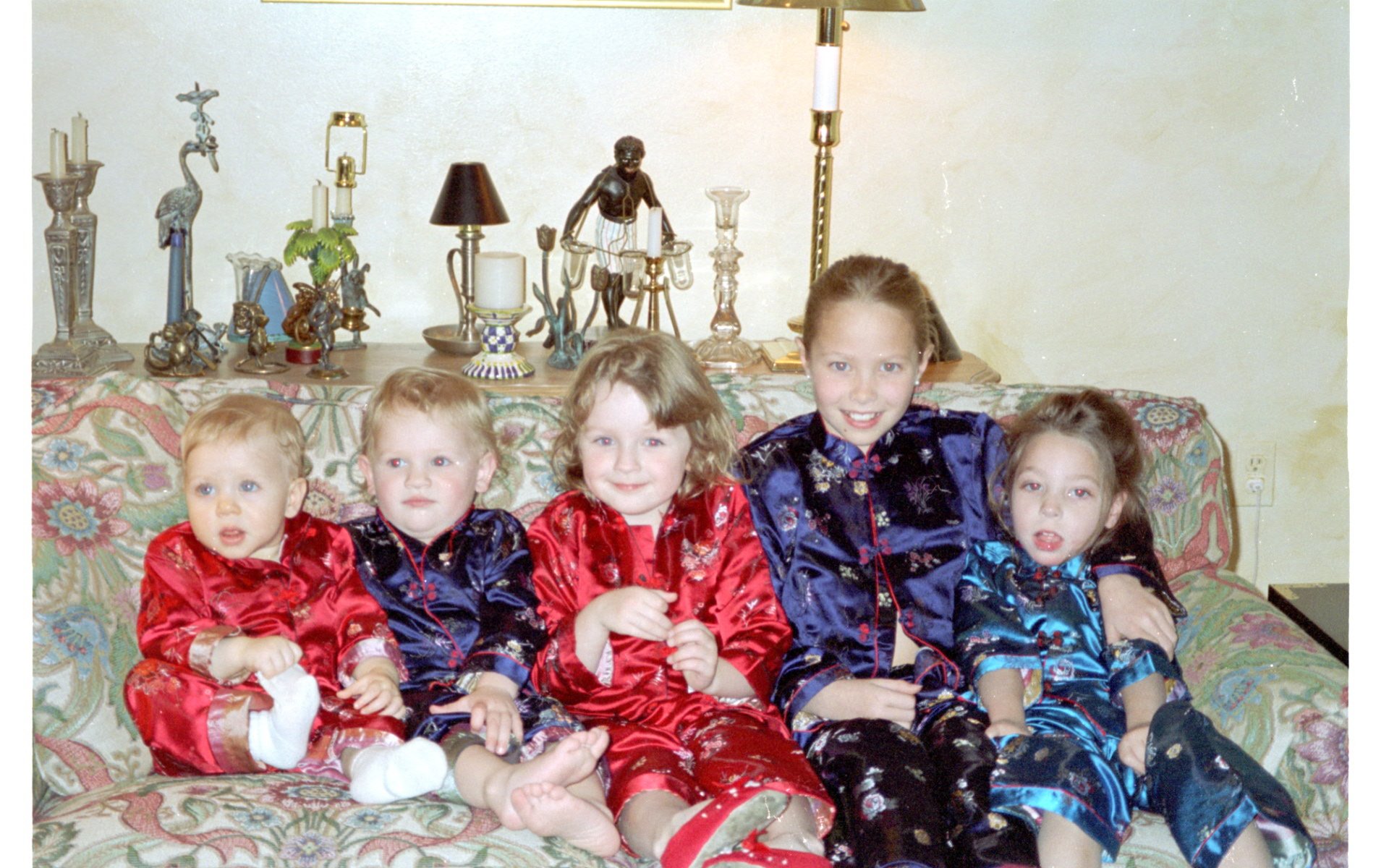 Six children sitting on a floral-patterned couch wearing matching colorful silk pajamas, with a beige wall and a wooden shelf with decorative items in the background.
