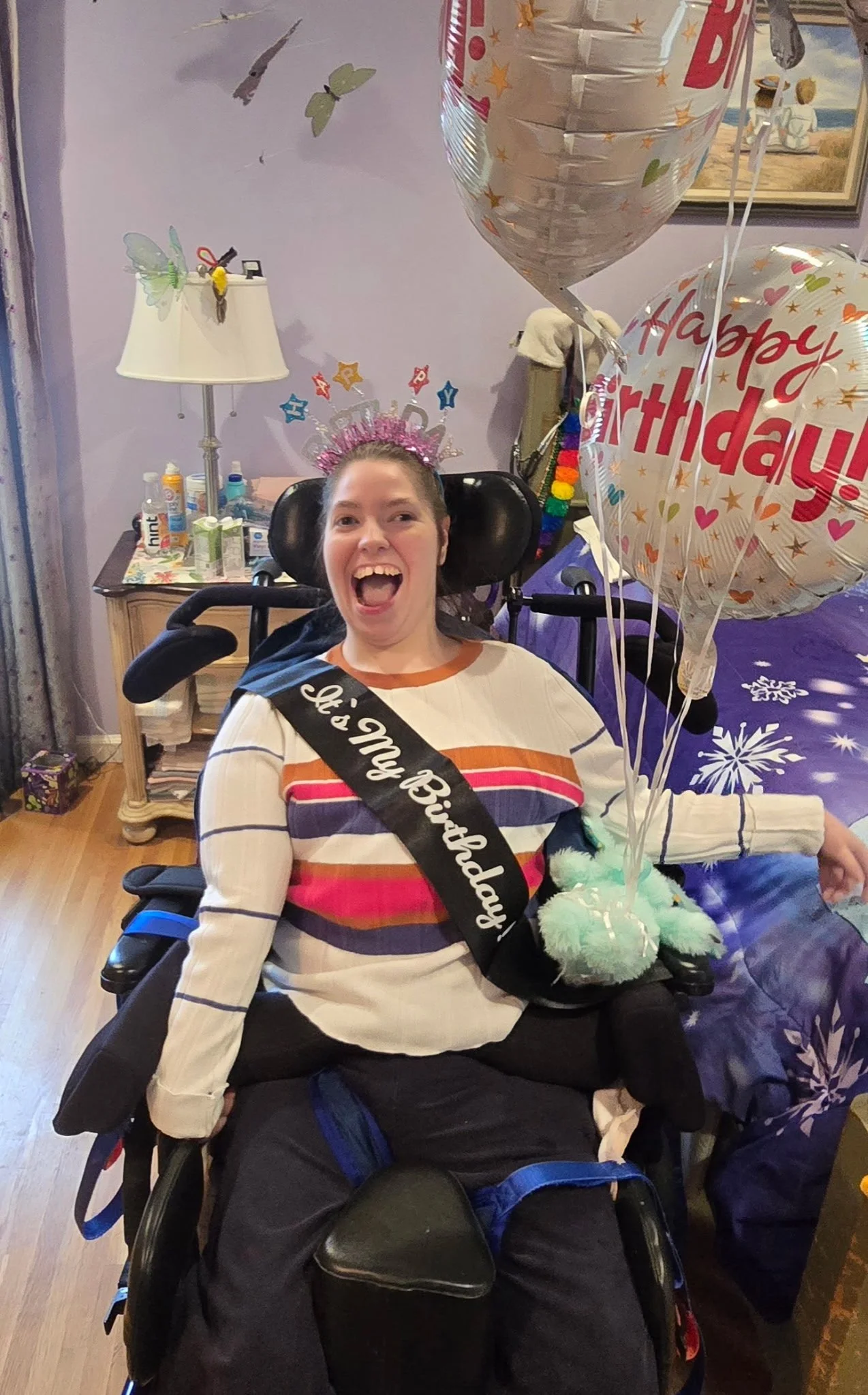 Woman celebrating birthday in a wheelchair, wearing a crown and a sash that reads 'It's My Birthday', with birthday balloons and decorations in the background.