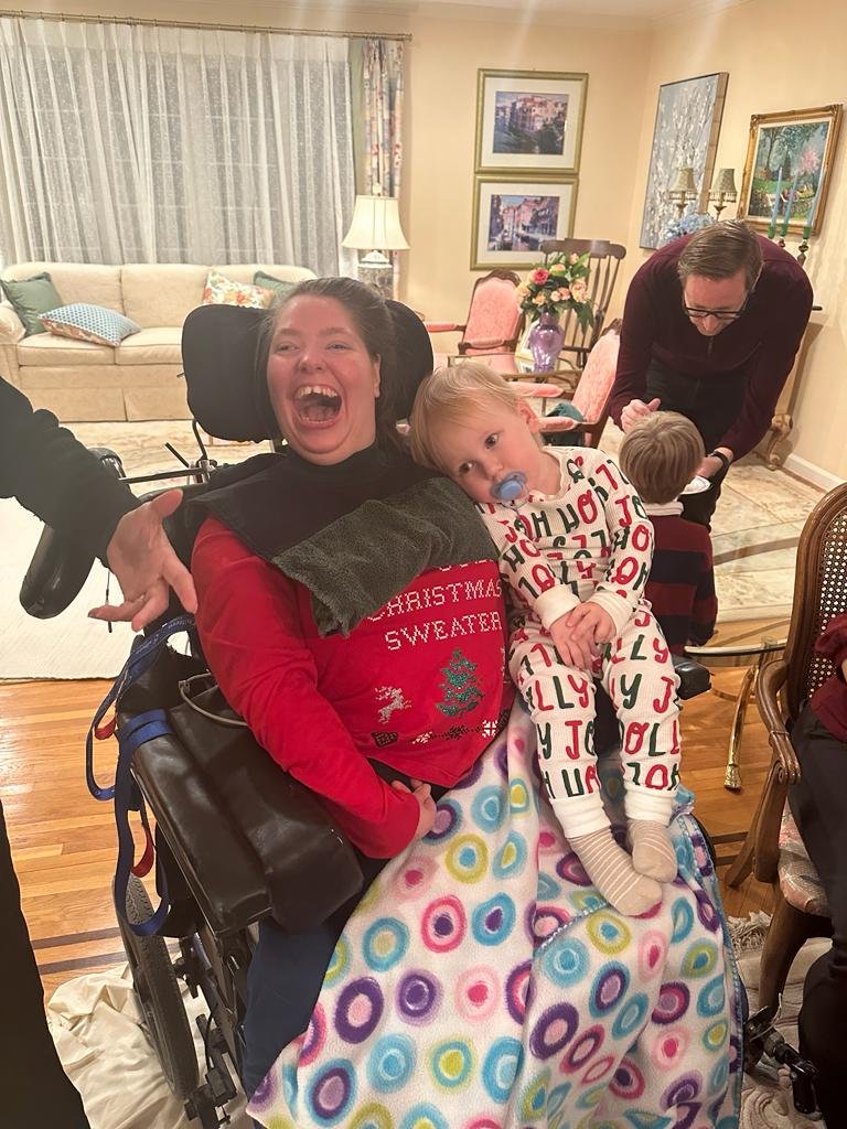 A young woman smiling joyfully in a wheelchair with a colorful blanket over her lap, holding a young girl sitting on her lap with a pacifier. A man is in the background, leaning over and talking to a small boy, in a cozy living room decorated with framed pictures, flowers, and lamps.