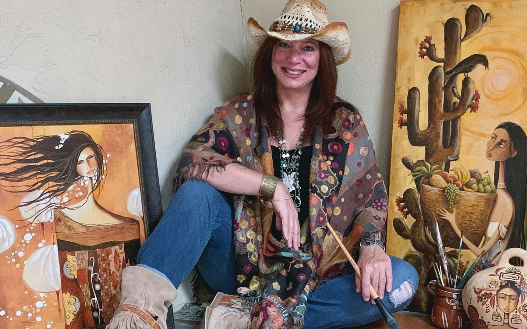 A woman seated on the floor next to two colorful paintings, wearing a cowboy hat, a patterned scarf, jewelry, jeans, and boots, surrounded by art supplies.