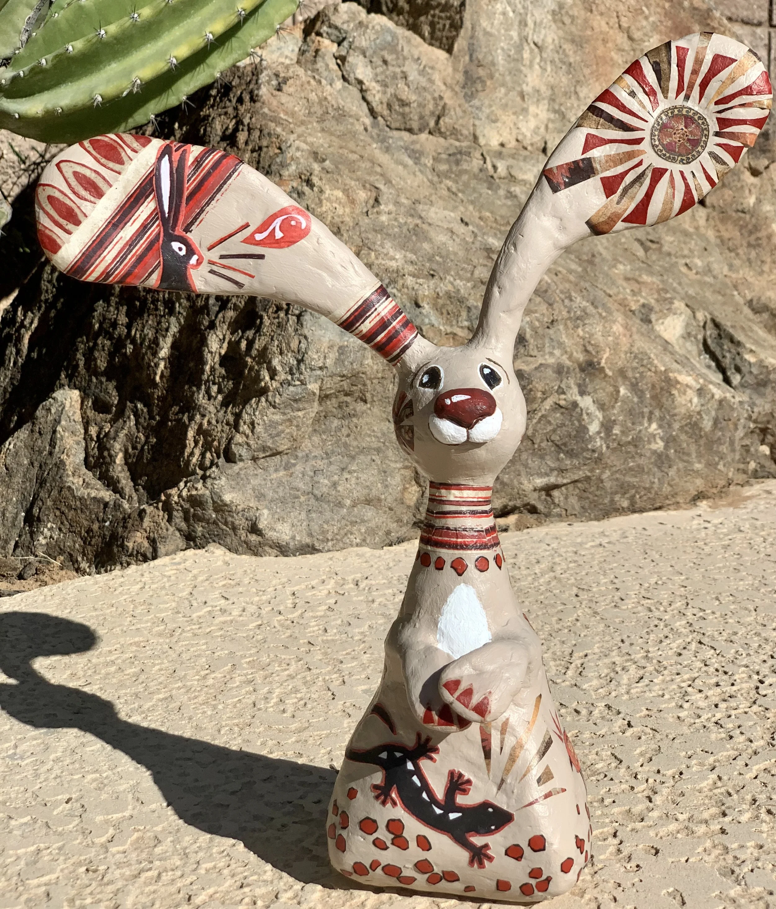 A decorative rabbit sculpture painted with native southwestern motifs and designs, featuring large ears with colorful patterns and a small lizard painted on the body. It is placed outdoors on a sandy surface near rocks and a cactus.