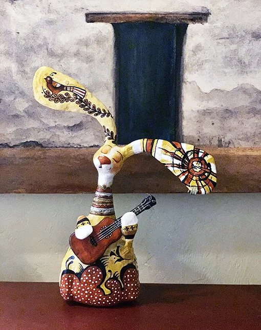 A decorative ceramic sculpture of a snake playing a guitar, with the upright part of the snake extending vertically. The sculpture is colorful with patterns, and the background shows a brick and wall.