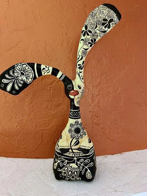 Decorative figurine of a dog with long ears, painted in black and white floral patterns, with a red nose, placed against an orange wall on a white surface.