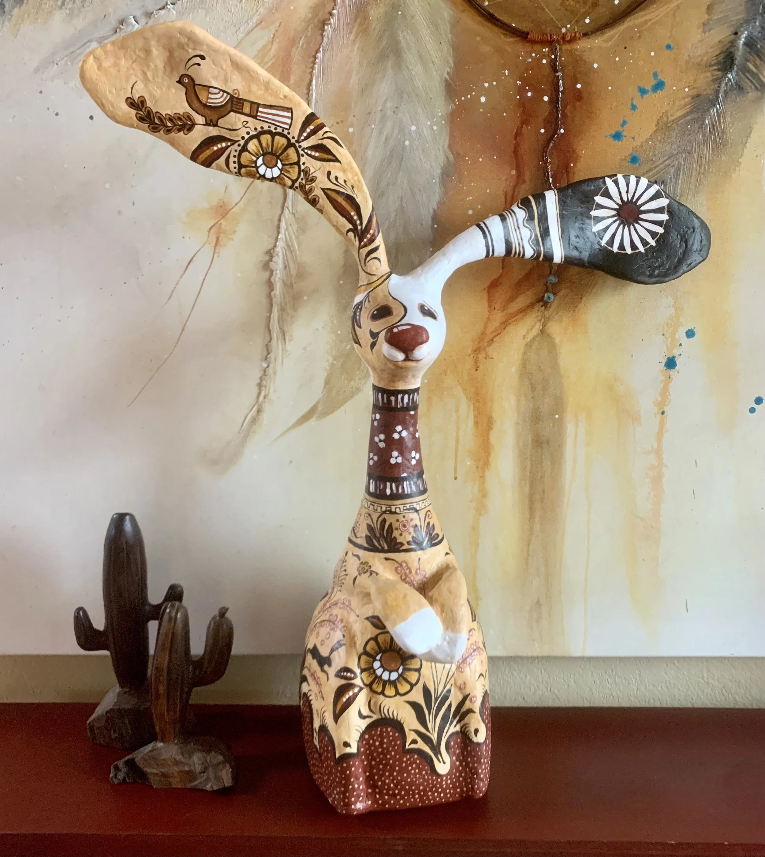 Decorative ceramic sculpture of a goat with whimsical painted designs, standing on a shelf next to a small wooden cactus figurine, with abstract colorful artwork in the background.