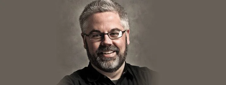 Portrait of a smiling man with glasses, gray hair, and a beard, wearing a black shirt against a gray background.