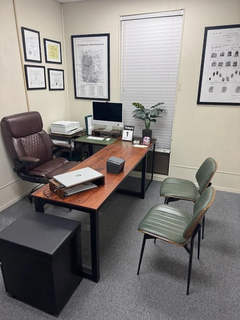 Office with a wooden desk, brown leather chair, green chairs, potted plant, computer, printer, and framed certificates on the wall.