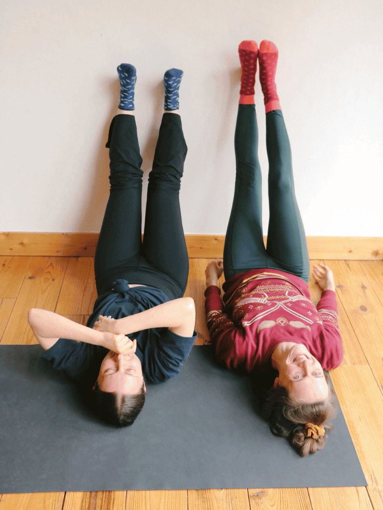 Two women are lying on their backs on yoga mats, with their legs raised against the wall. One woman is wearing a navy shirt and camouflage socks, while the other is in a maroon shirt with strange socks with dots.