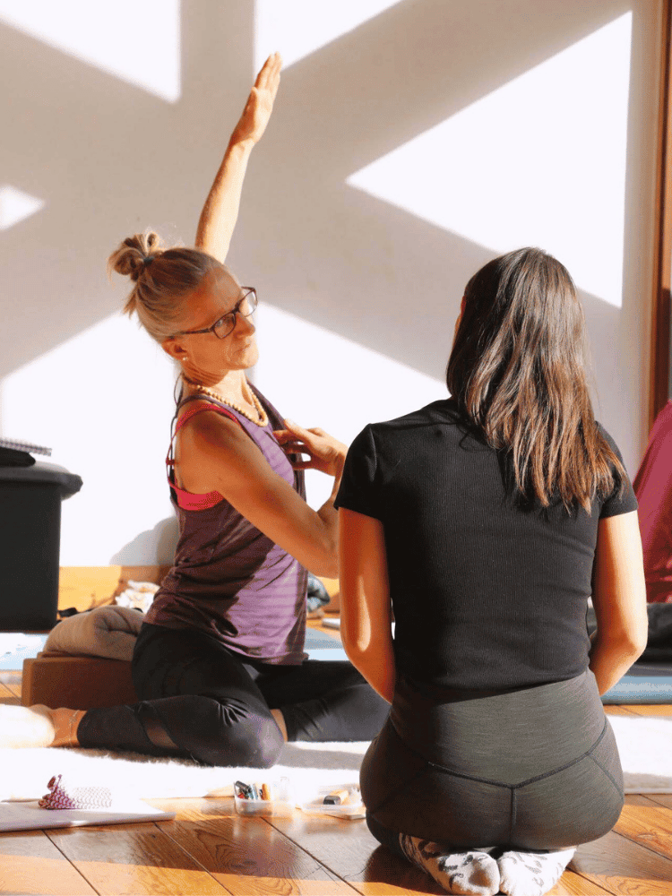A woman in a purple tank top and black leggings sits on a beige mat, stretching her right arm up while another woman with long dark hair, wearing a black top and gray leggings, sits facing her on a wooden floor. The sunlight creates shadows on the wall behind them.