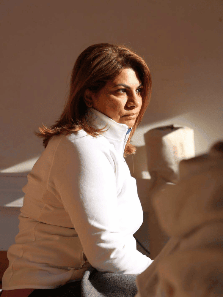 A woman with brown hair wearing a white zip-up jacket, sitting indoors with a relaxed expression, in front of a blurred background.