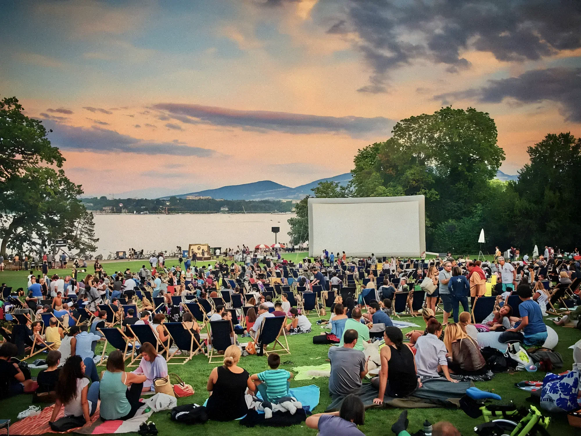 Crowd watching outdoor movie on a large inflatable screen by a lake Geneva at sunset, with trees and mountains in the background.
