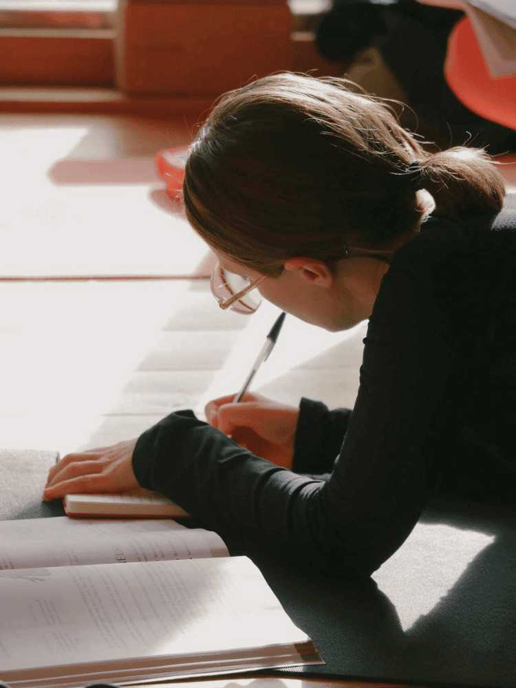 A person with glasses and brown hair tied in a ponytail, sitting on the floor and writing in a notebook with sunlight coming through a window.
