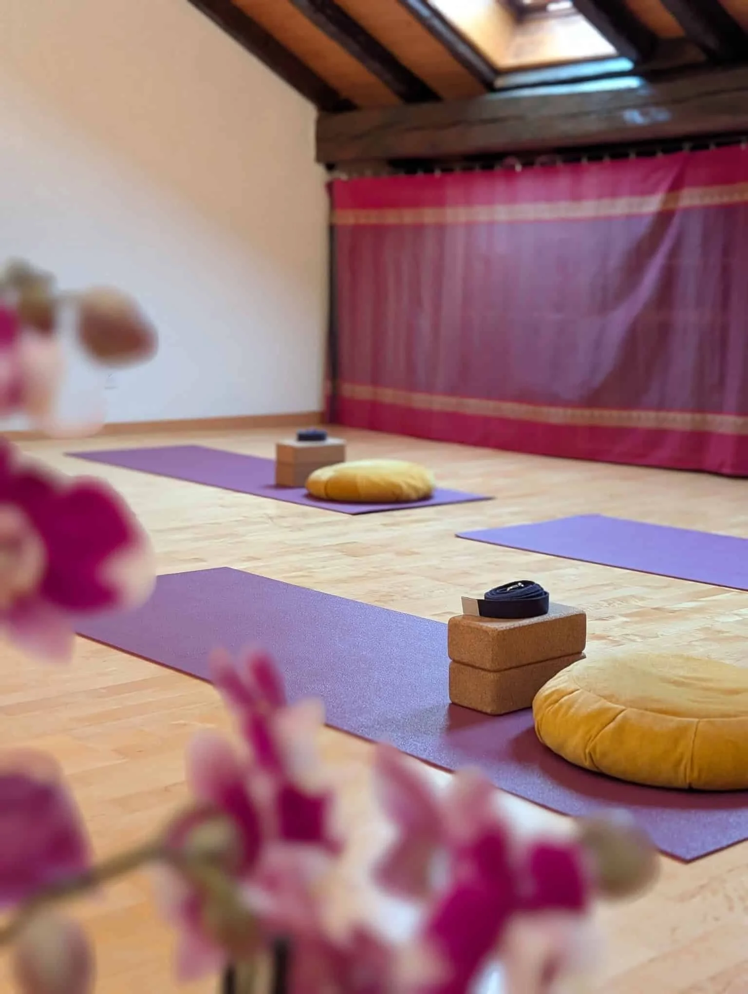 Yoga mats, cushions, and blocks arranged on a wooden floor in a cozy room with a pink curtain and wooden ceiling.