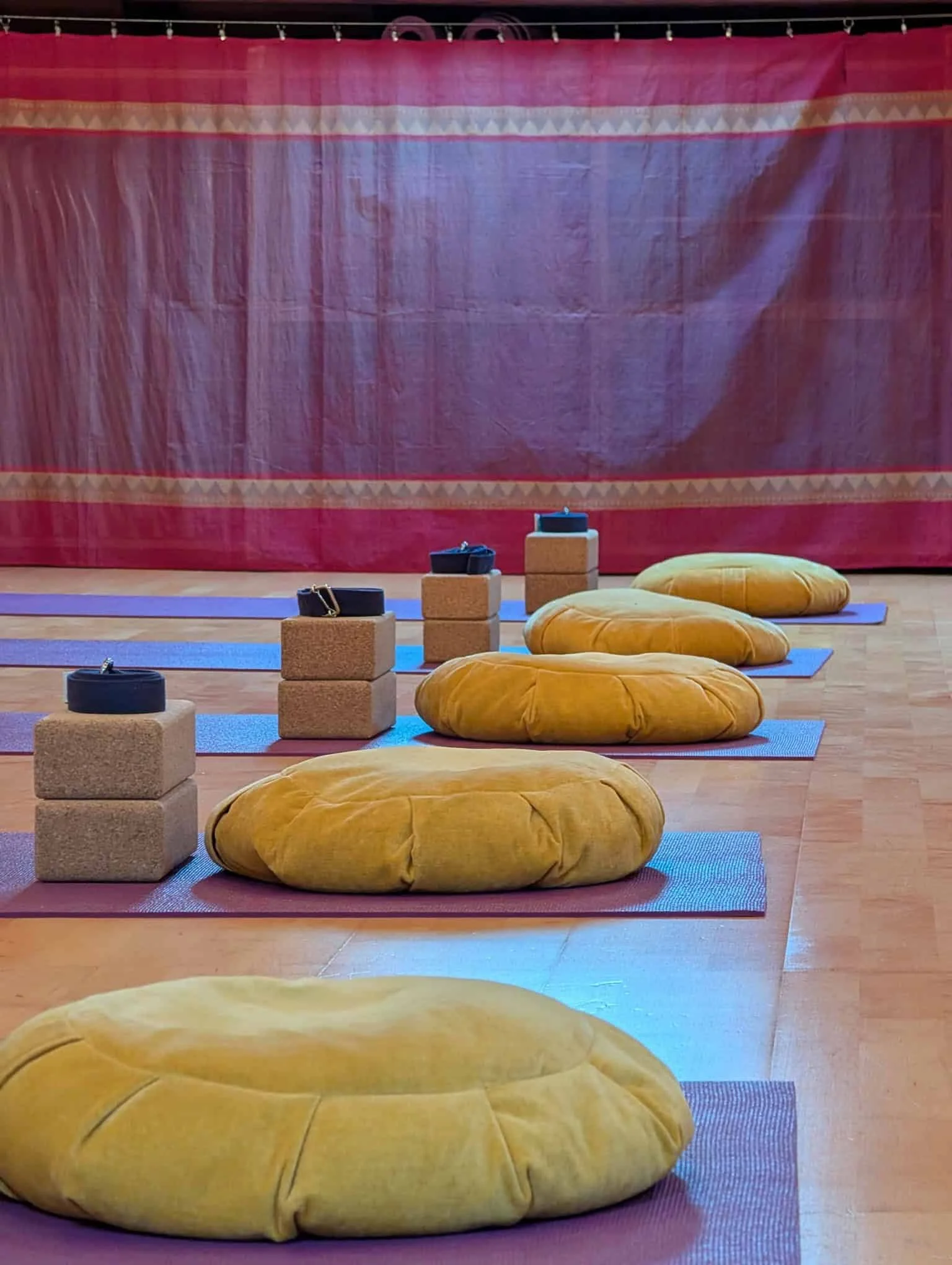 Yoga or meditation studio setup with yellow cushions, purple yoga mats, and stacked cork blocks with straps, against a pink curtain backdrop.
