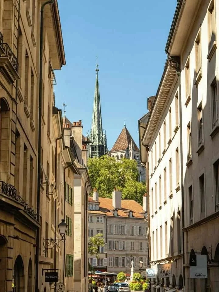 A narrow street in Geneva old town with tall buildings on either side, a church with a tall spire in the background, and outdoor cafes with umbrellas in the foreground.