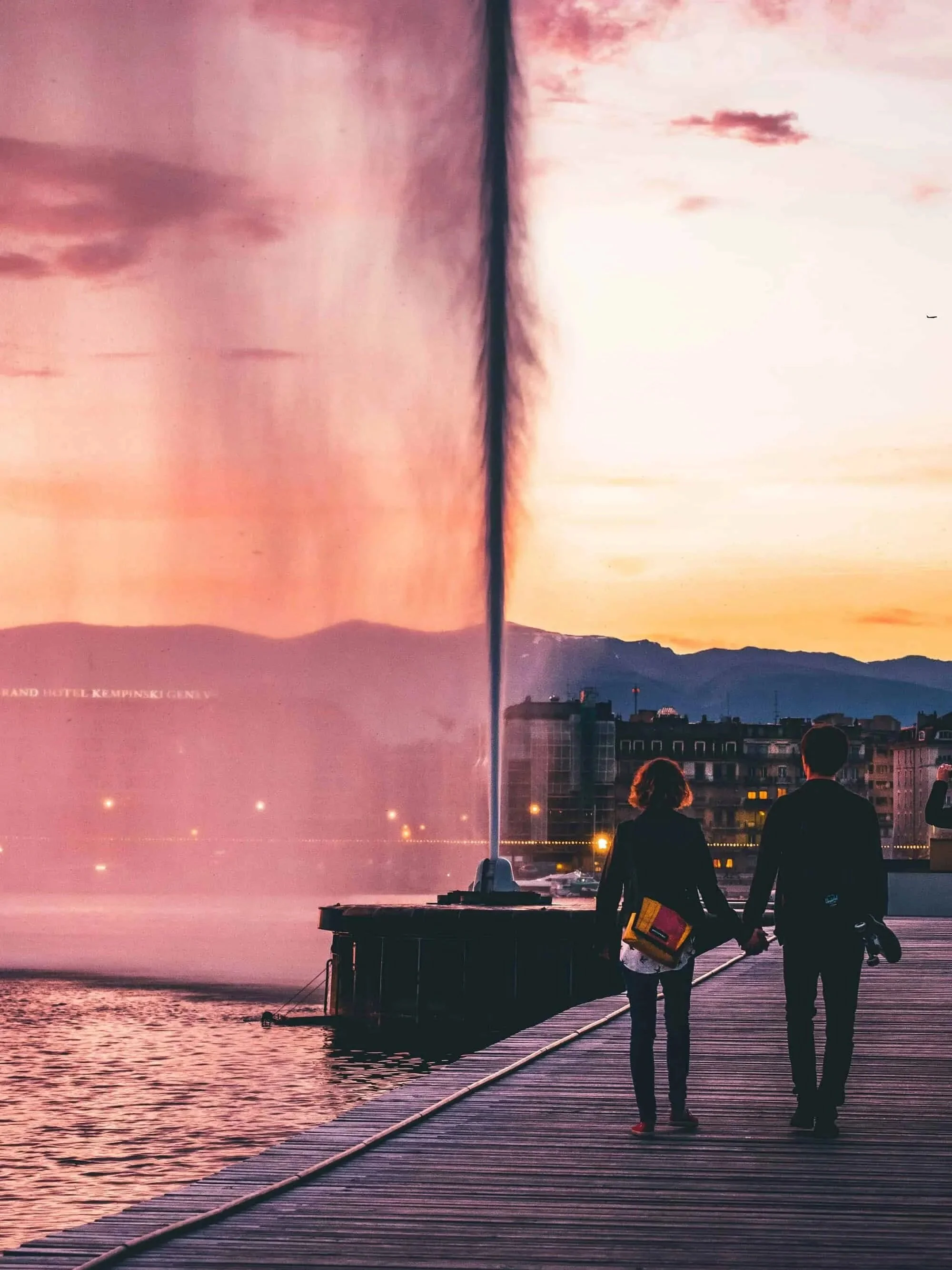 A couple walking hand in hand along a wooden waterfront promenade at sunset, with a cityscape and mountains in the background, and a fountain jetting water into the air.
