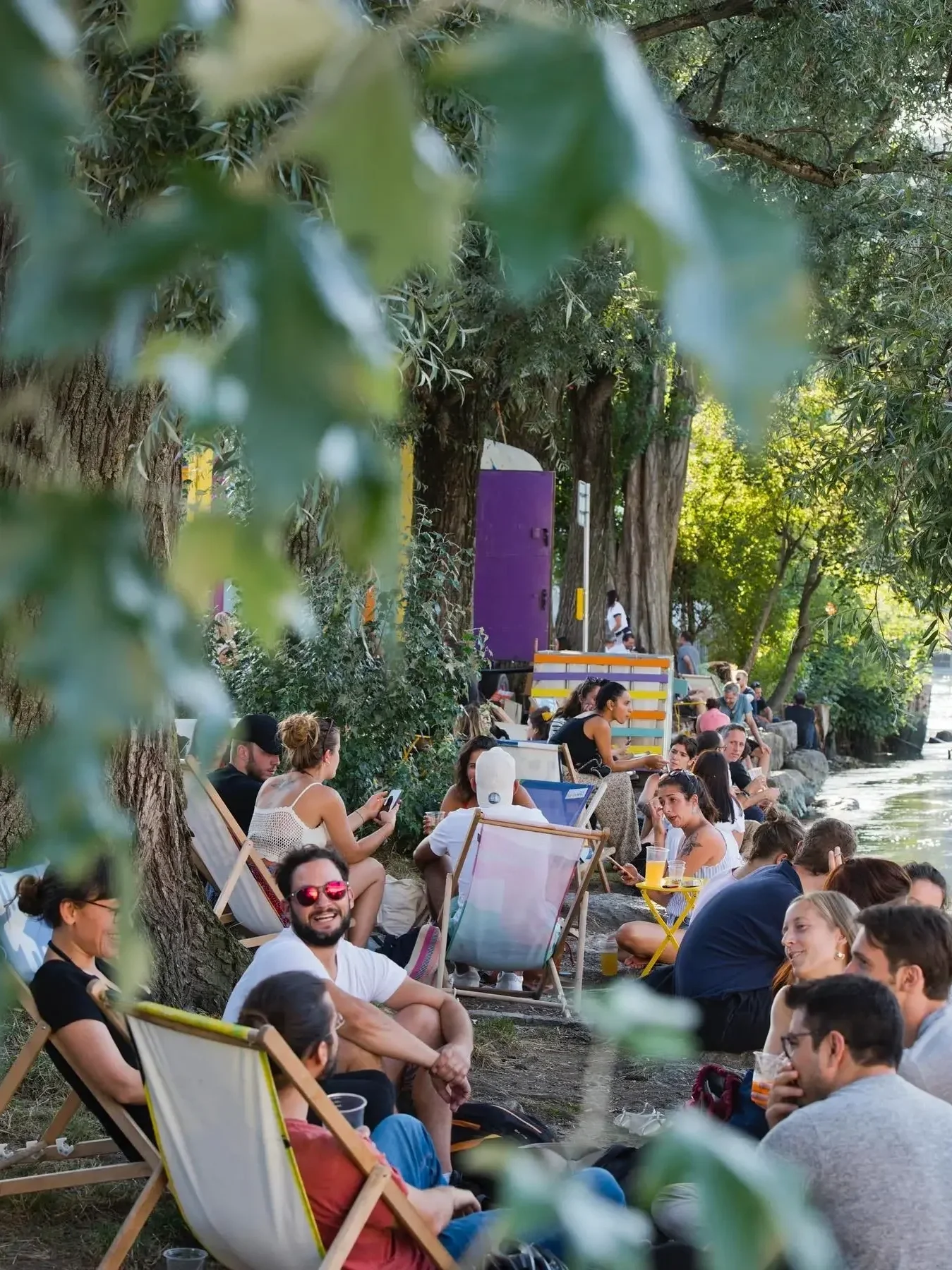 People sitting on the ground and in chairs along a riverbank in Geneva, socializing and enjoying drinks in a sunny outdoor setting surrounded by trees.