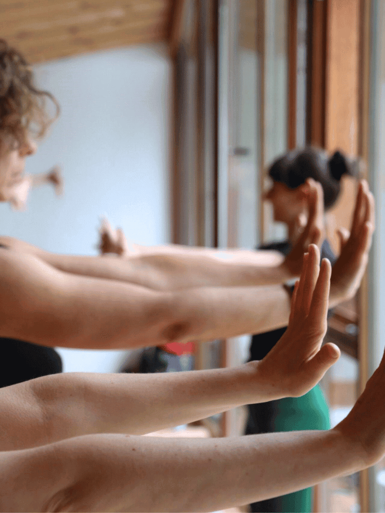 People practicing yoga or meditation with arms extended and palms facing outward near a large window.