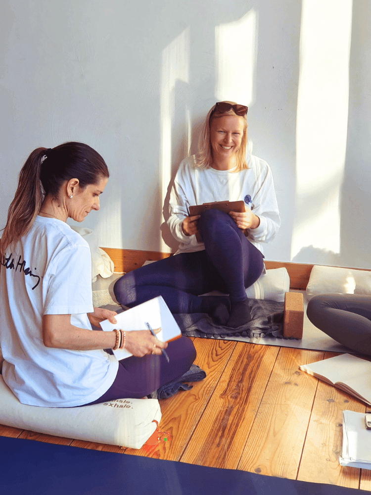 Two women sitting on a wooden floor, smiling and taking notes, with sunlight coming through a window behind them.