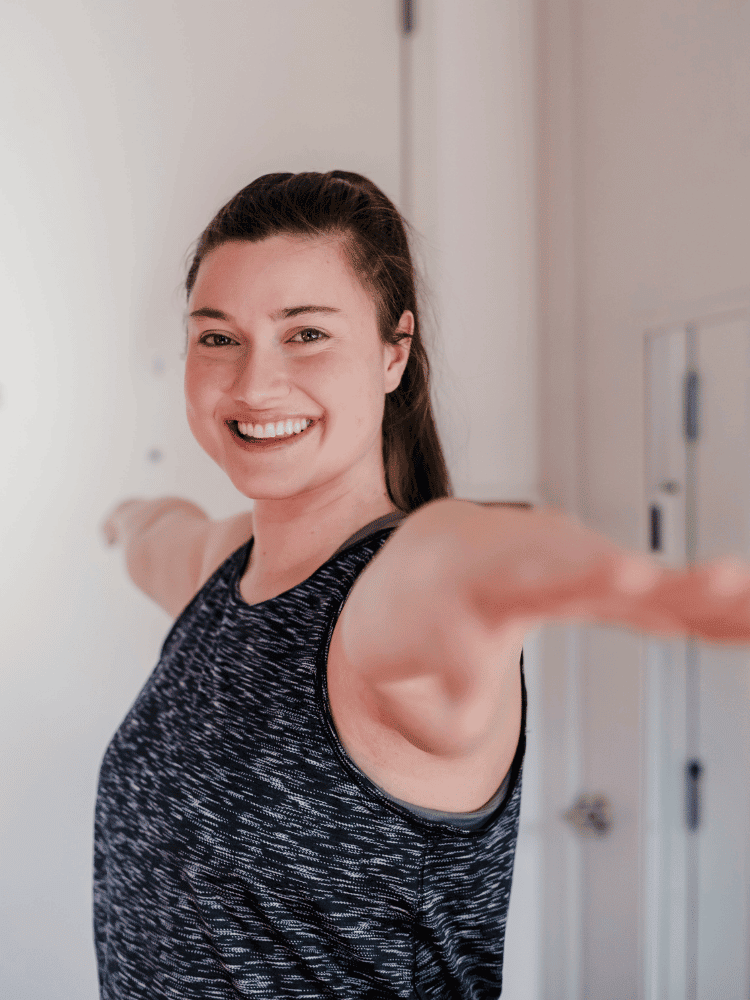 Woman with brown hair in a ponytail smiling and reaching out with arms extended in Warrior 2 yoga pose.