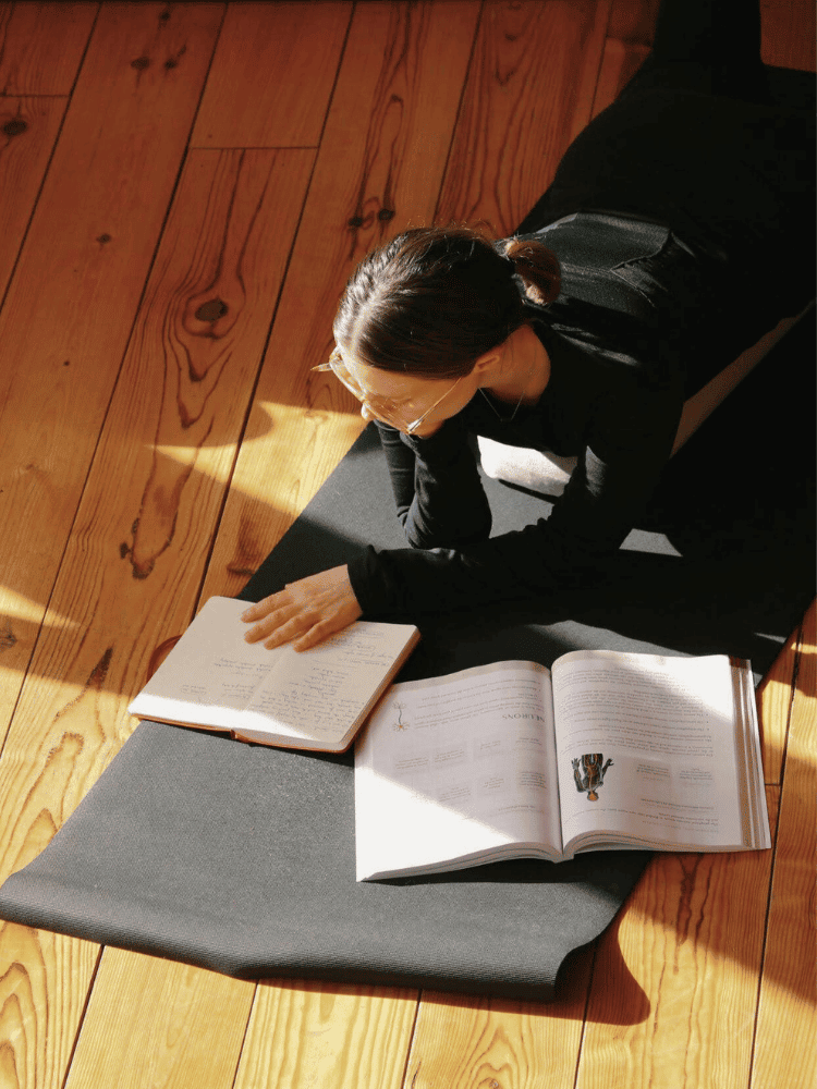 A woman lying on a yoga mat on wooden floor, reading a notebook and a YTT textbook, with sunlight casting shadows.