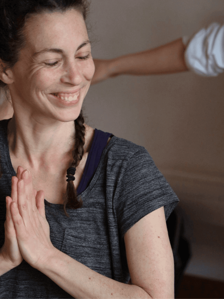 A woman smiling with closed eyes, hands pressed together in a prayer position, wearing a dark gray athletic shirt, with a braid over her shoulder.