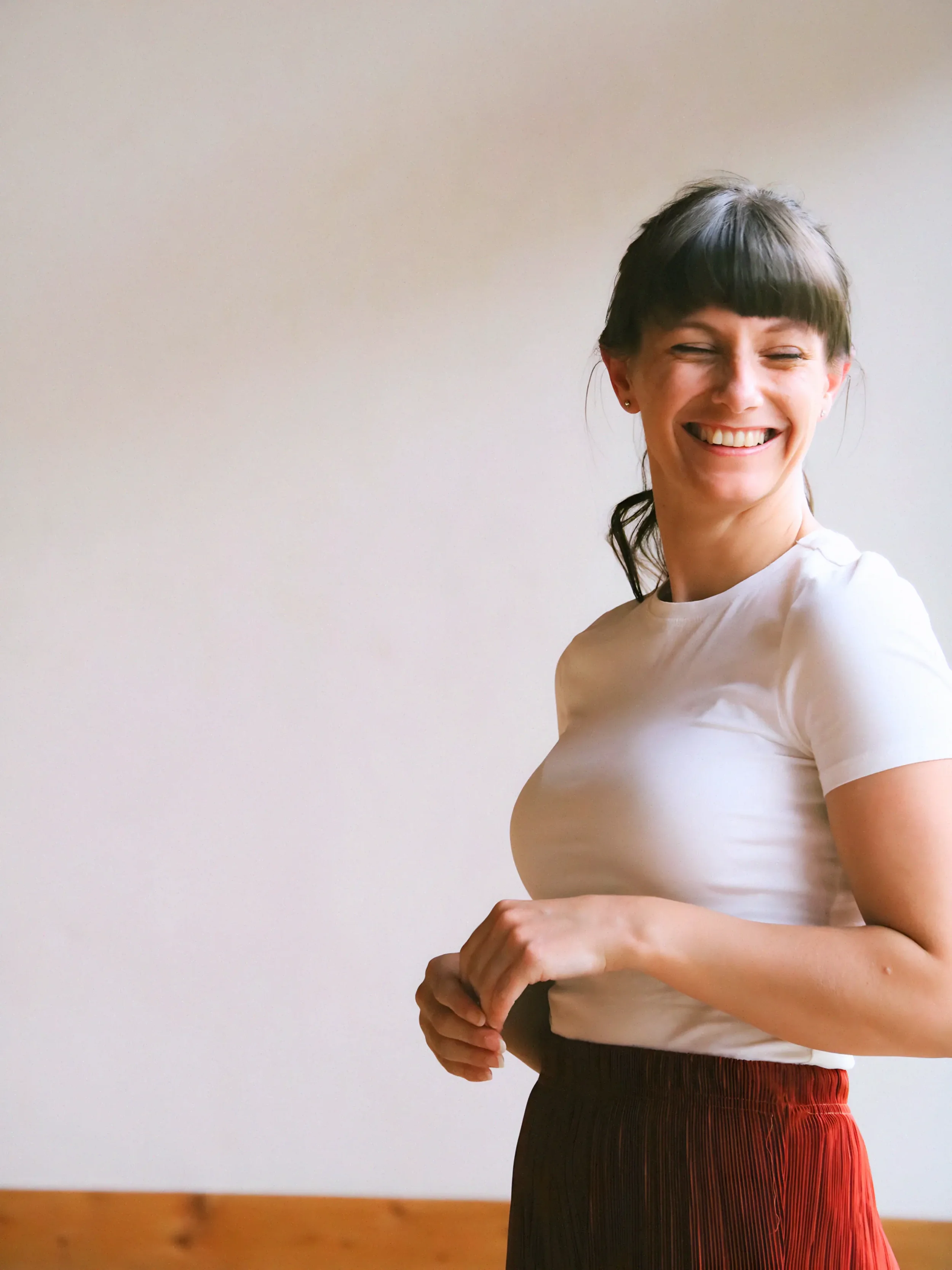 A woman with dark brown hair and bangs, wearing a white t-shirt and a dark red pleated skirt, smiling and looking to her right, standing against a plain light-colored wall.