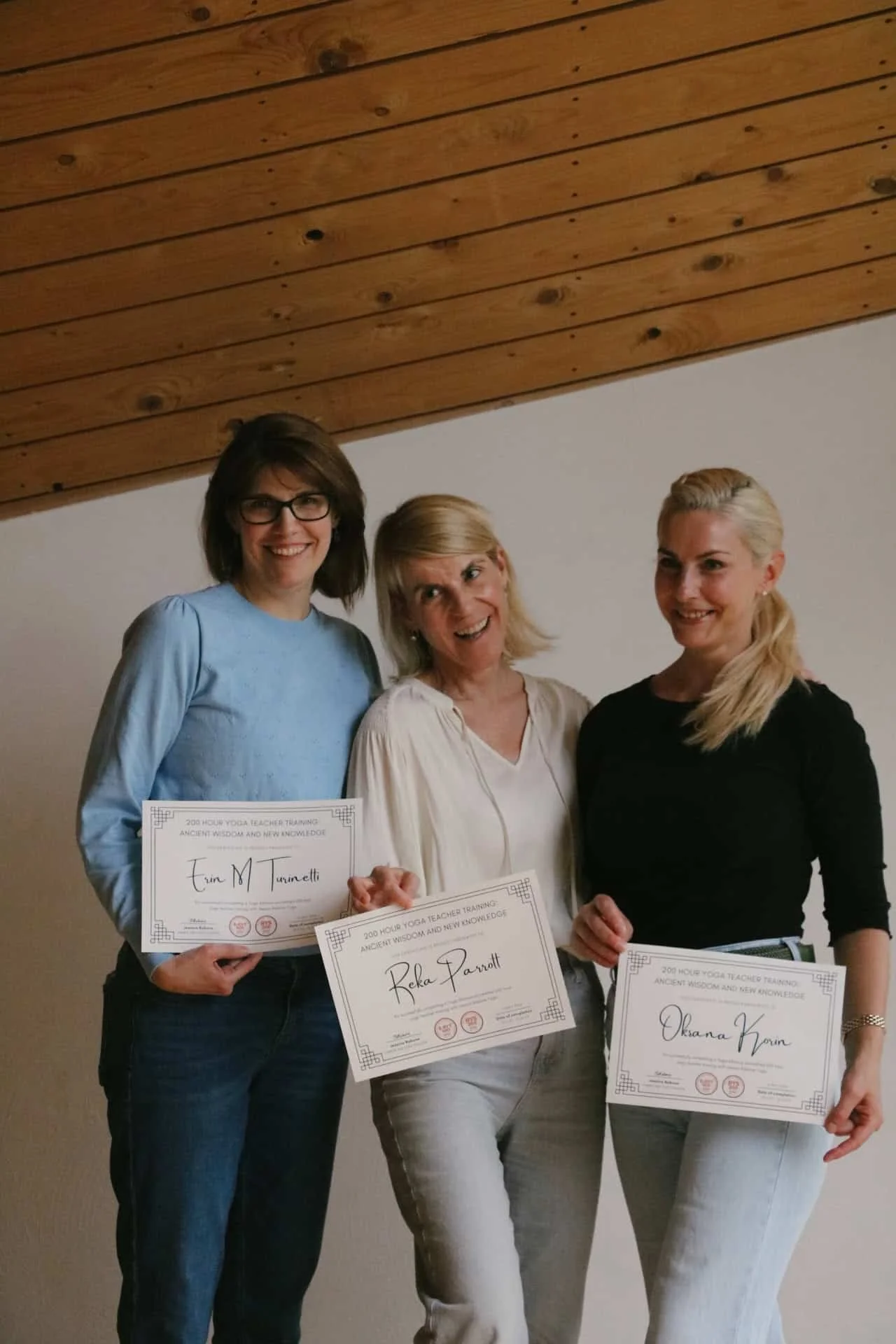 Three women standing together, holding Yoga Alliance YTT certificates, smiling indoors in front of a plain wall and wooden ceiling.