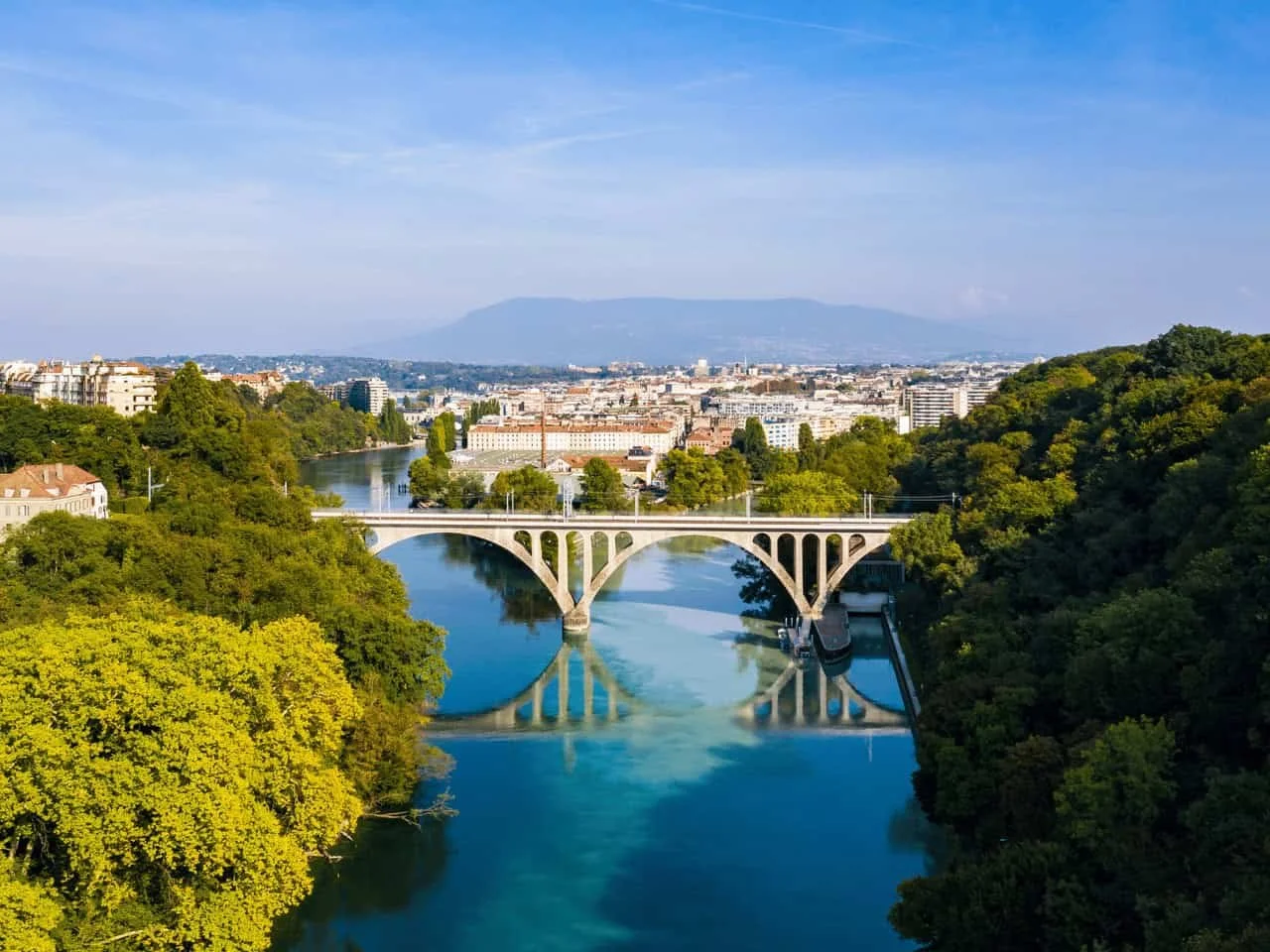 A scenic view of Geneva with a river flowing through it, a stone bridge crossing the river, surrounded by green trees and urban buildings in the distance, with mountains in the background under a blue sky.