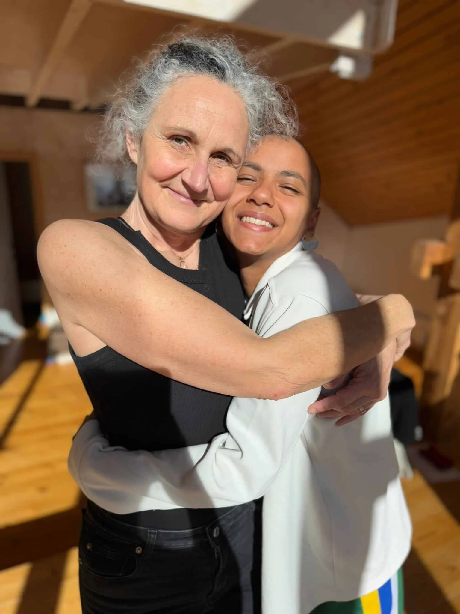 An elderly woman and a young lady hugging, both smiling warmly at the camera, indoors with wooden walls and ceiling.